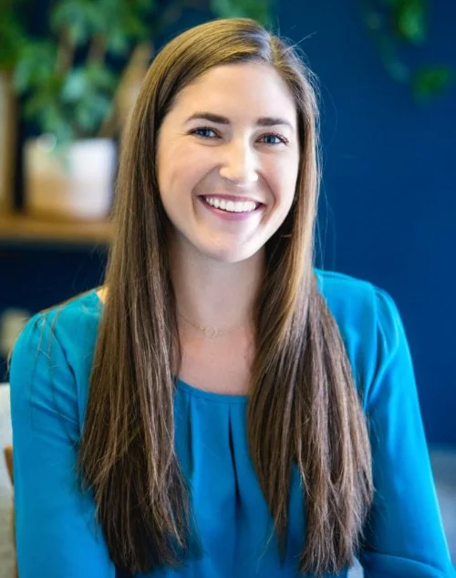 A young woman with long brown hair smiling, wearing a blue shirt, sitting indoors with a plant and decorative items in the background.