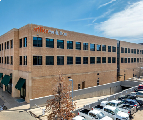 A large office building with a sign reading 'office evolution' on the top corner. The building has multiple windows, a parking lot with several cars, and a clear blue sky above.