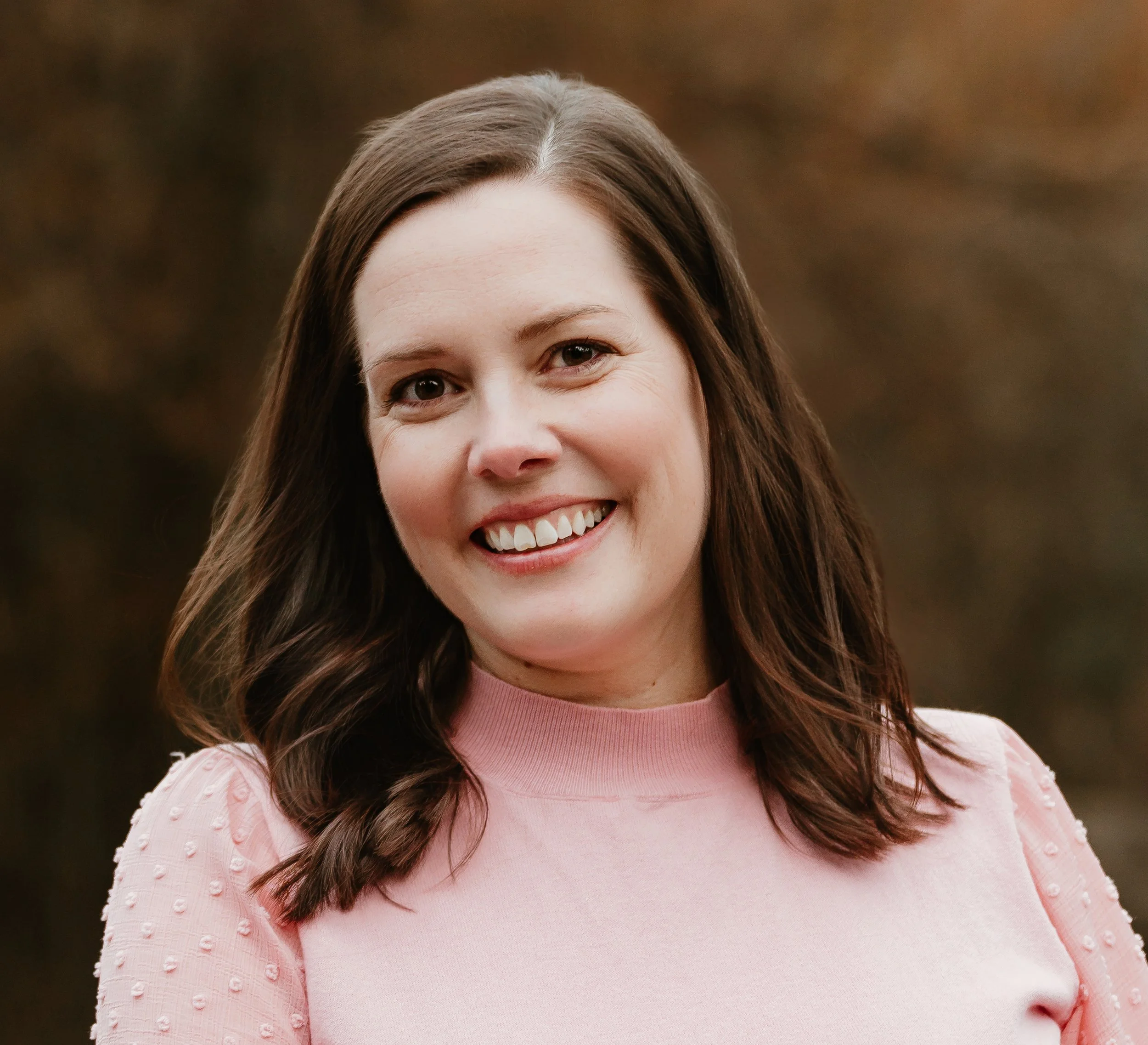 Yolanda, a young woman with shoulder-length brown hair, smiling, wearing a purple hoodie and a gold necklace with a circular pendant, outdoors with blurred trees and grass in the background.