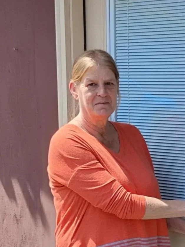 A middle-aged woman with light brown hari pulled back stands in front of a door with blinds.