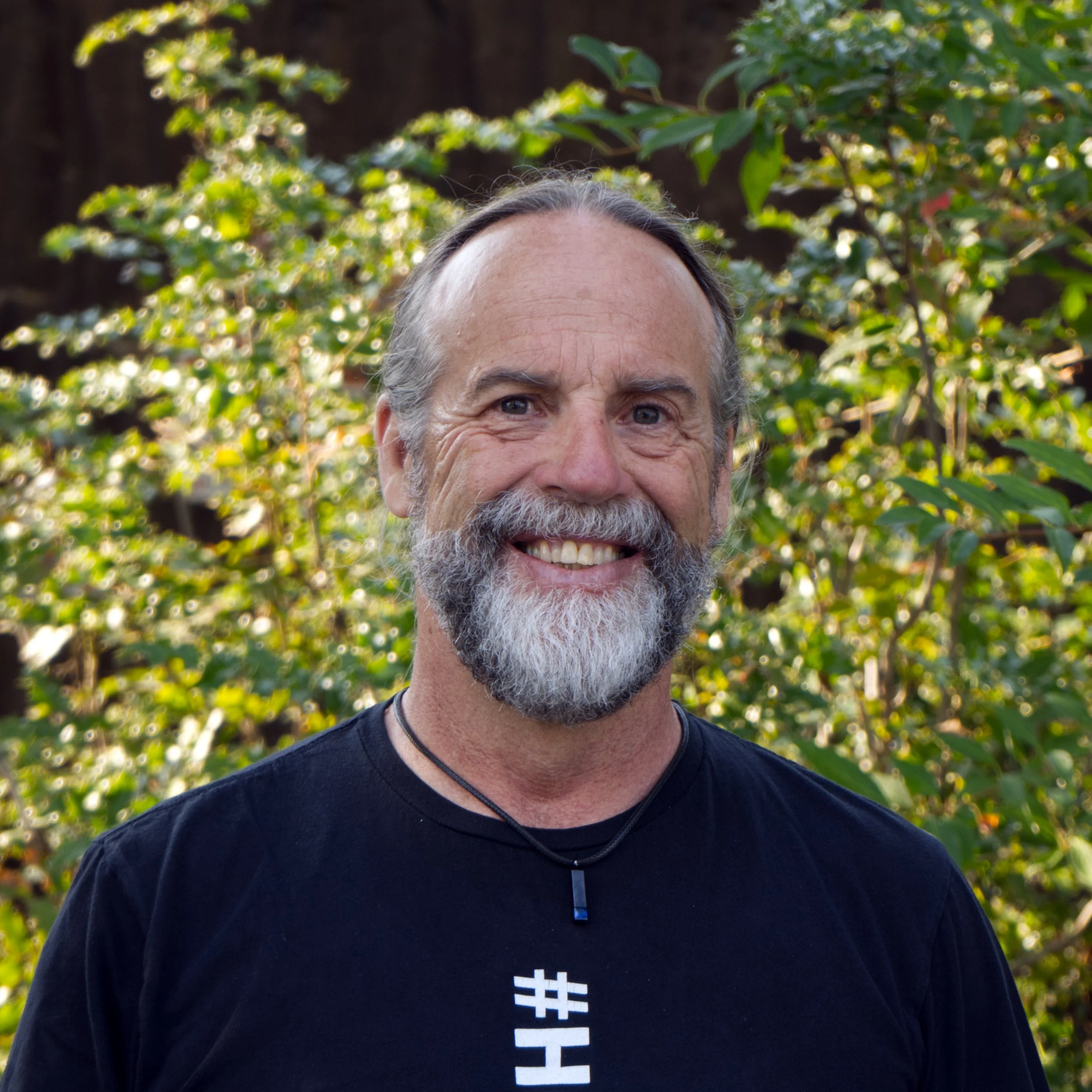 Mark, a smiling middle-aged man with a gray beard and mustache wearing a black T-shirt and a black necklace standing outdoors with green foliage in the background.