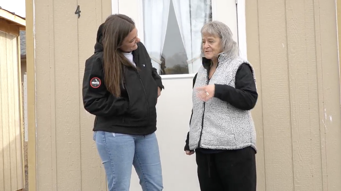 Case manager Cait, a young woman with long brown hair wearing a black jacket and blue jeans, smiles at Josephine, a short gray-haired woman in black pants, a black shirt, and a fuzzy gray vest, as they stand in a front of a microshelter.