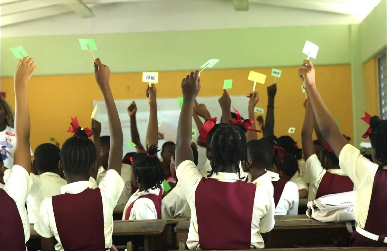 Students participating in our Myths vs. Realities Activity as Part of the ‘Break the Stigma: Pass the Pad’ Safe Cycle School Tour. Image of a Classroom of students from the back, with their hands in the air holding 'TRUE' or 'FALSE' cards