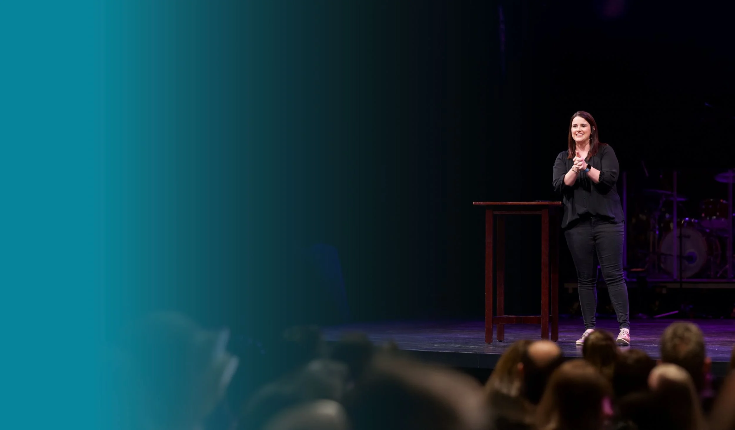 A woman stands on a stage speaking into a microphone, next to a small podium, with a dimly lit audience in the foreground and a gradient teal background on the left side of the image.