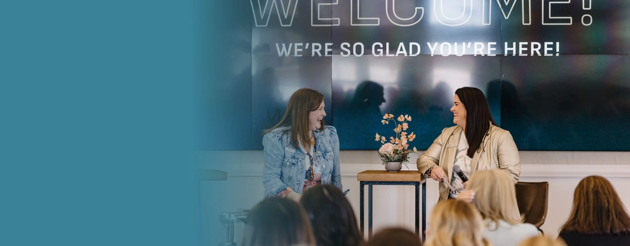 Two women sitting on stools and speaking to an audience. A small table between them holds a flower arrangement.