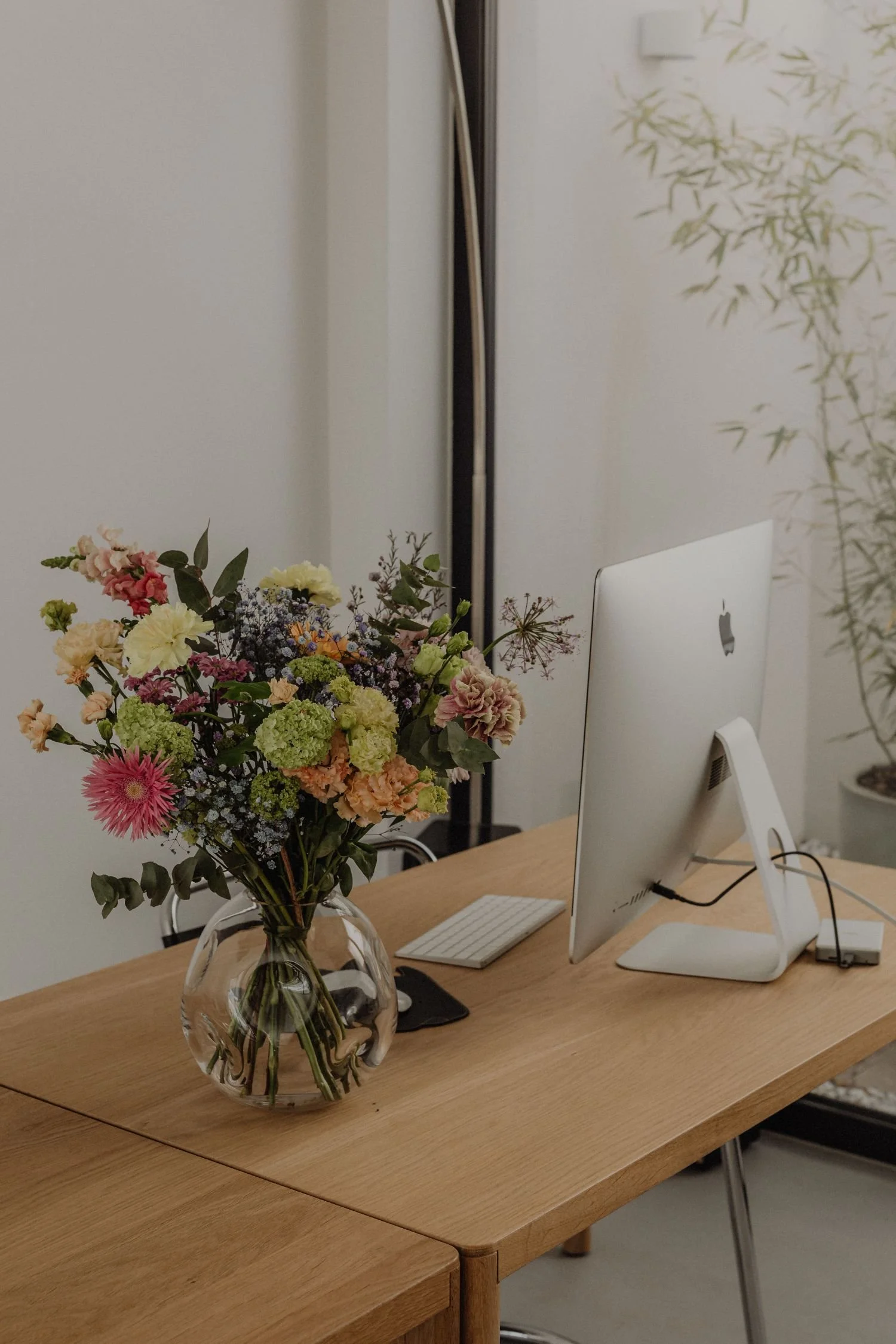 A sleek desk features an Apple computer and a vase of colorful flowers including pink, yellow, and white blooms, adding a touch of nature to the modern workspace. A bamboo plant is visible in the background.