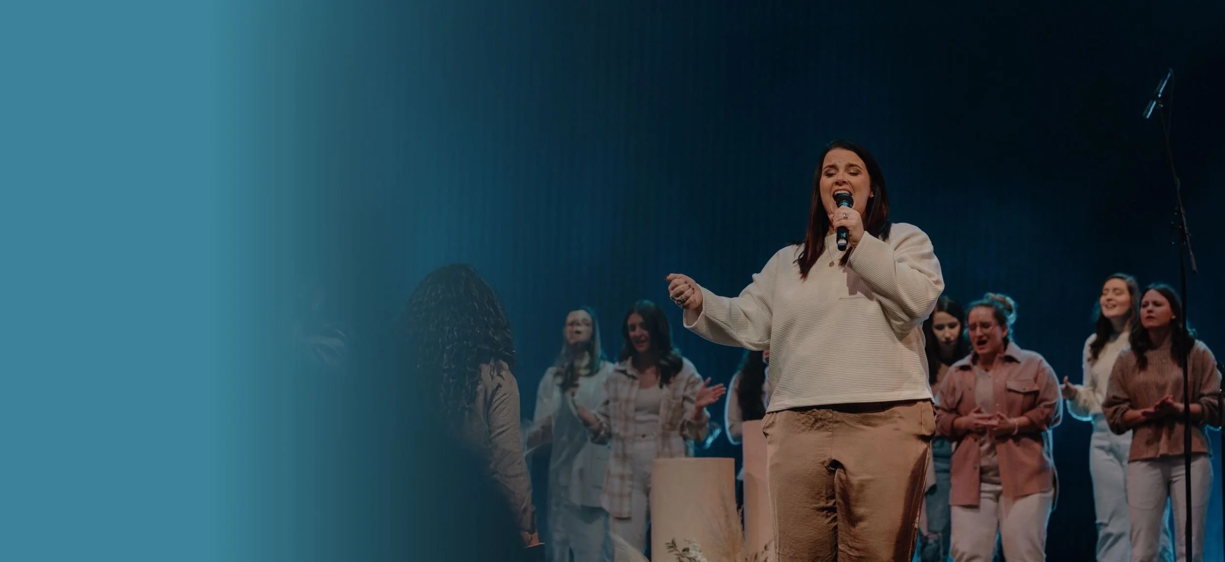 A woman is singing passionately into a microphone on stage, surrounded by a group of people in casual attire. The background is dark with a blue gradient on the left side of the image.