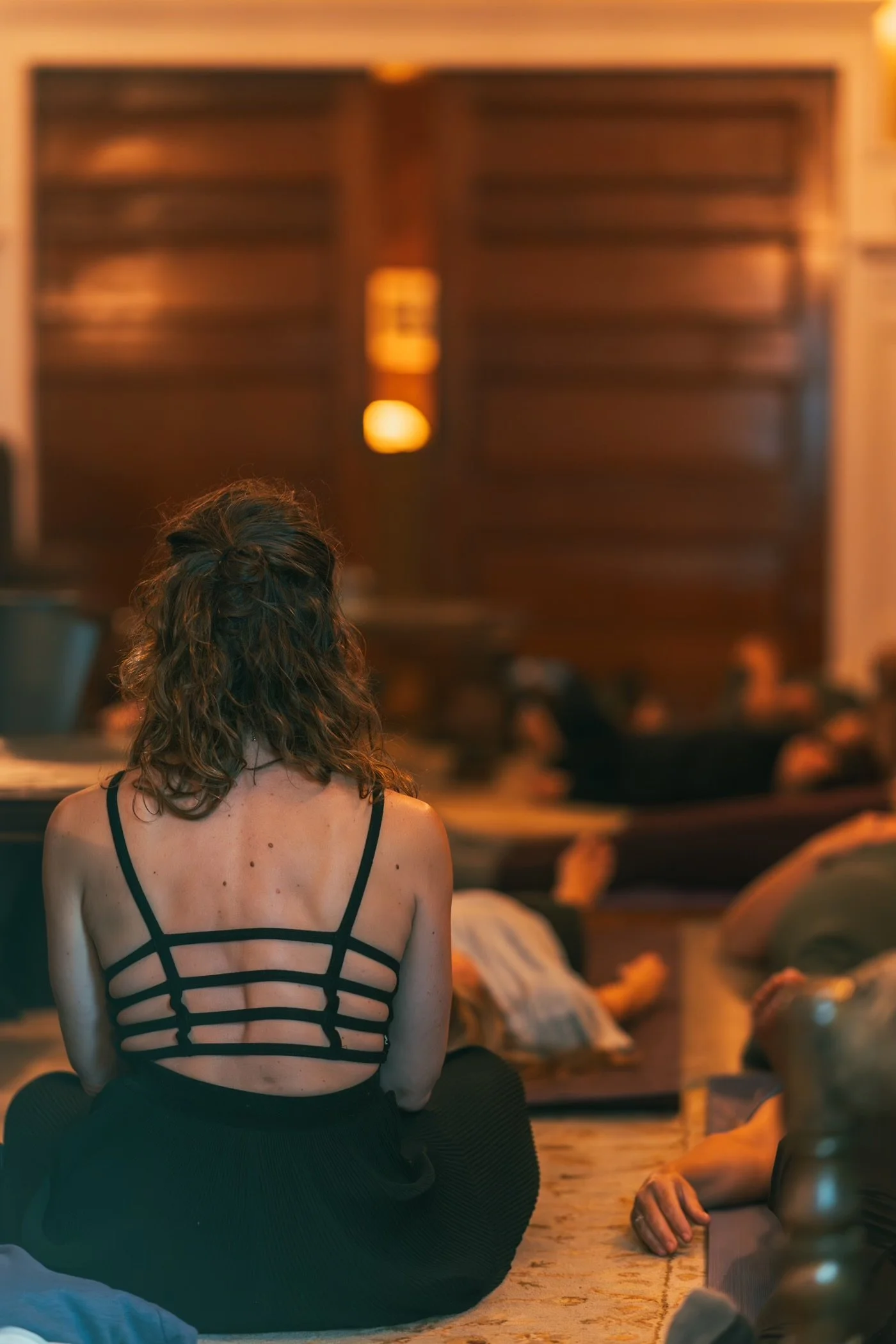 A woman with wavy brown hair sitting cross-legged on the floor, facing away, wearing a black strappy back top. In the background, other people are lying on the floor, some appearing to be resting or sleeping, in a warm and softly lit room.