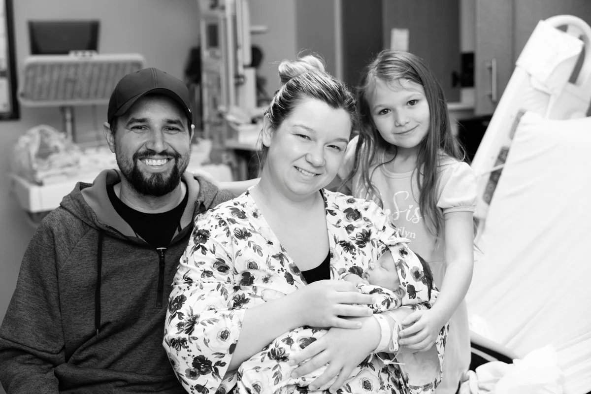 A black and white photo of a mother holding a newborn baby with her husband on the left and a little girl on the right. In the background is hospital equipment.