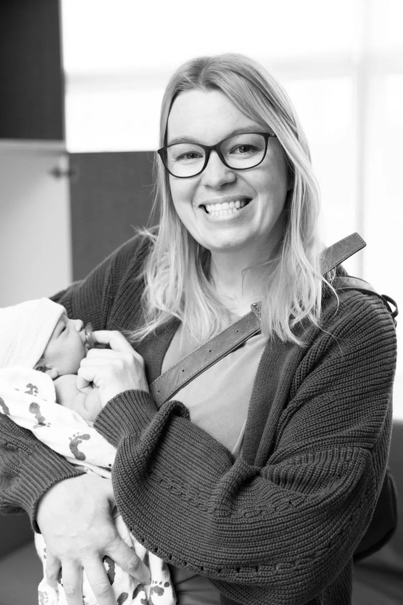 A black and white photo of a woman with blonde hair and glasses, smiling and holding a newborn baby with a hat and a pacifier in her mouth.