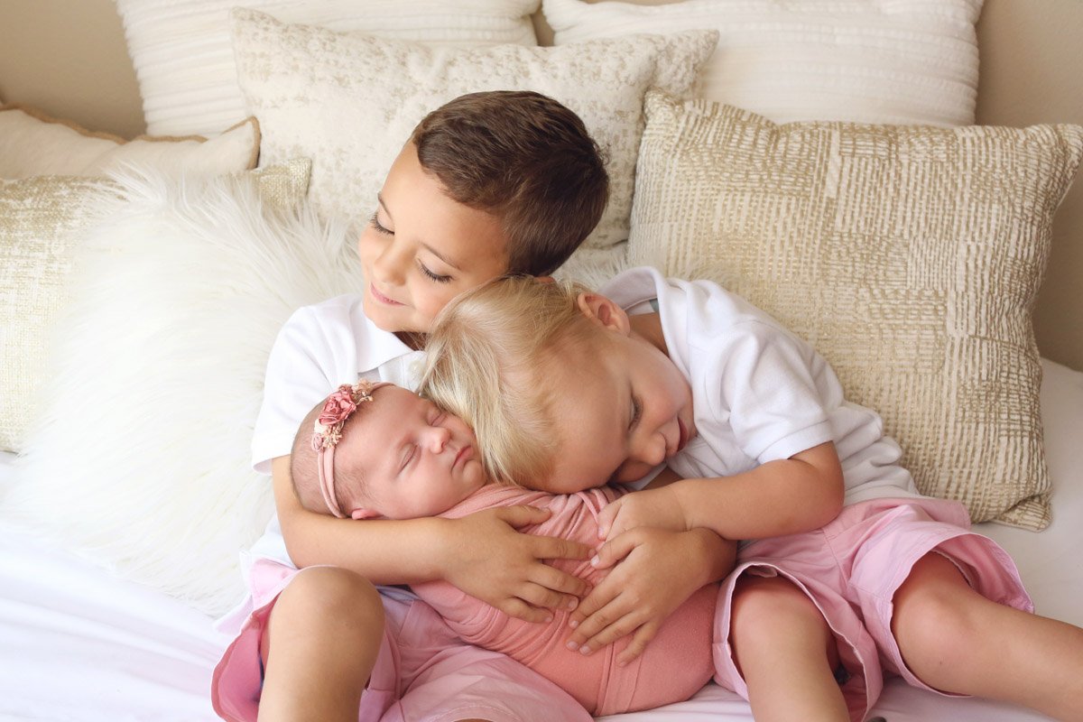 Photo of two young boys holding and hugging their newborn sister in pink with a flower headband. They are on a white bed with textured, neutral pillows behind them.