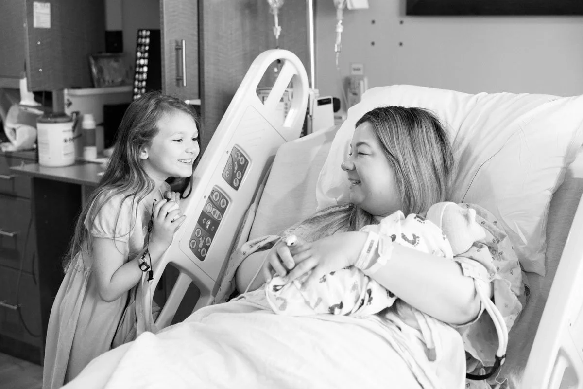 Black and white photo of a little girl smiling at the edge of a hospital bed, seeing her mom in a hospital bed holding a newborn. In the background are hospital cabinets and equipment.