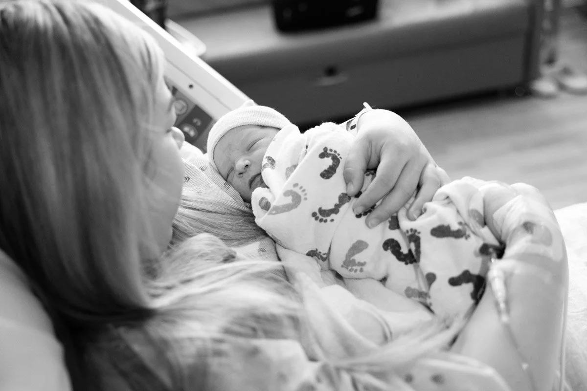 A black and white photo of a baby wrapped in a footprint-patterned blanket in the arms of a mother in a hospital bed.