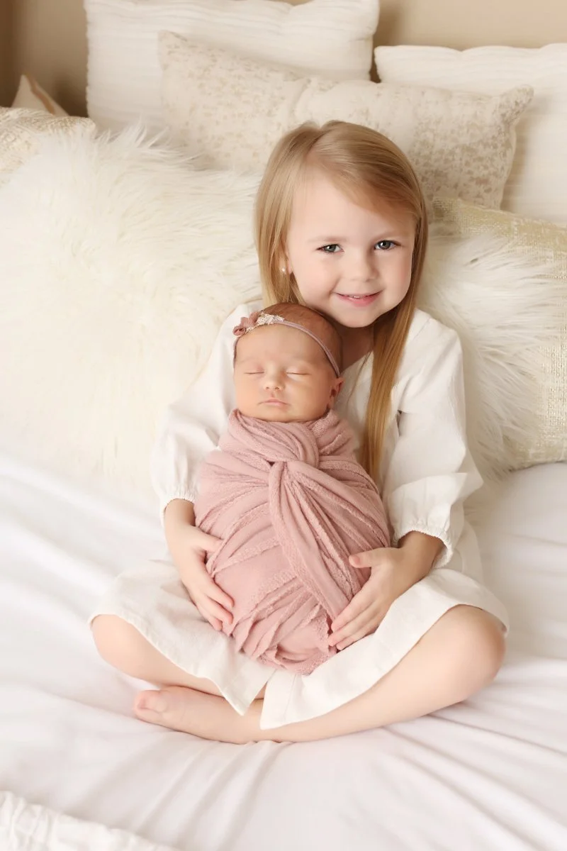 A young girl with long blonde hair, wearing a white dress and sitting on a bed holding a newborn wrapped in a dusty pink blanket. They are on a white bed with white, textured pillows behind them.
