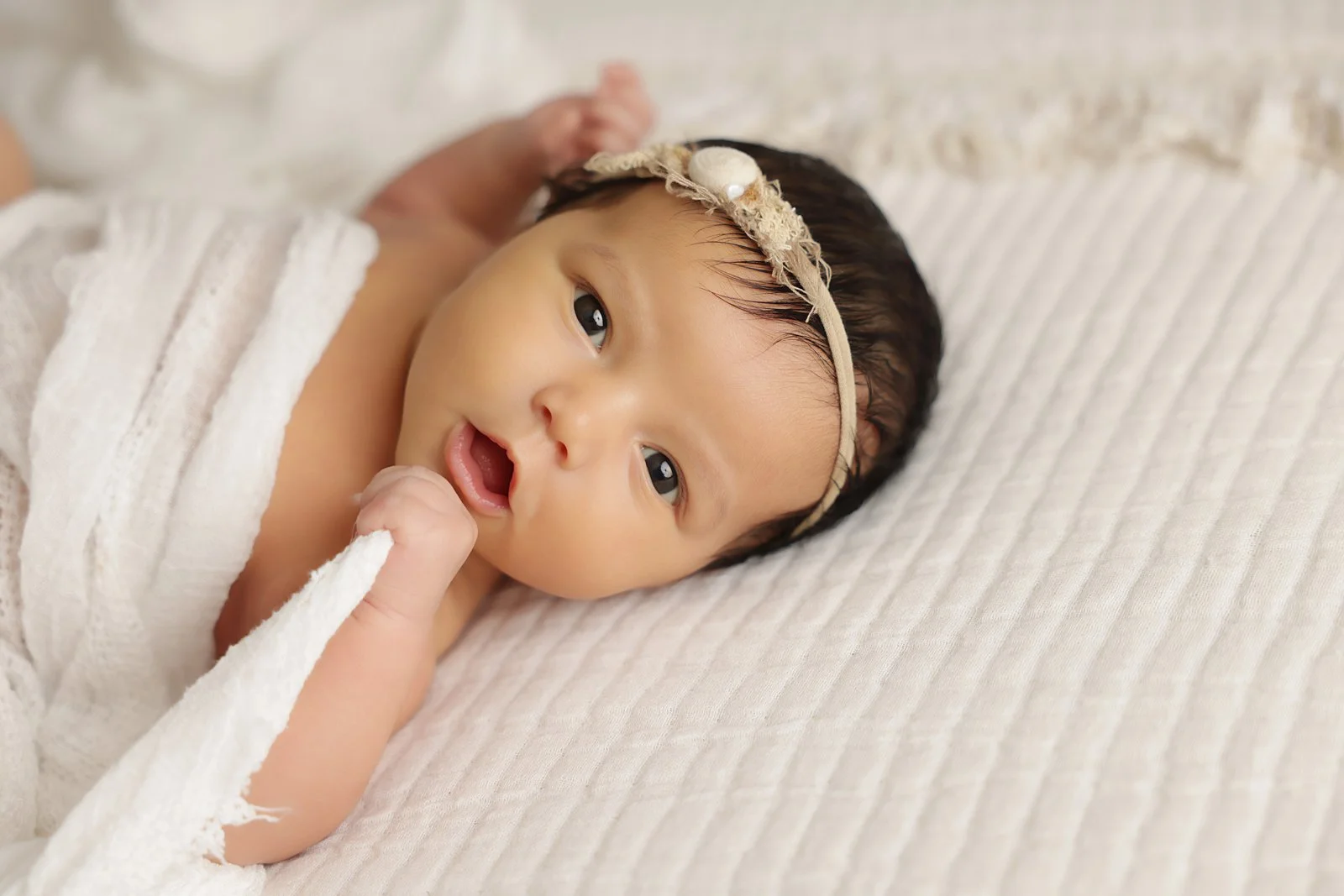 Photo of a newborn baby with dark brown hair and eyes, wearing a headband and looking at the camera. She is laying on her back under a white blanket on a white, textured surface.