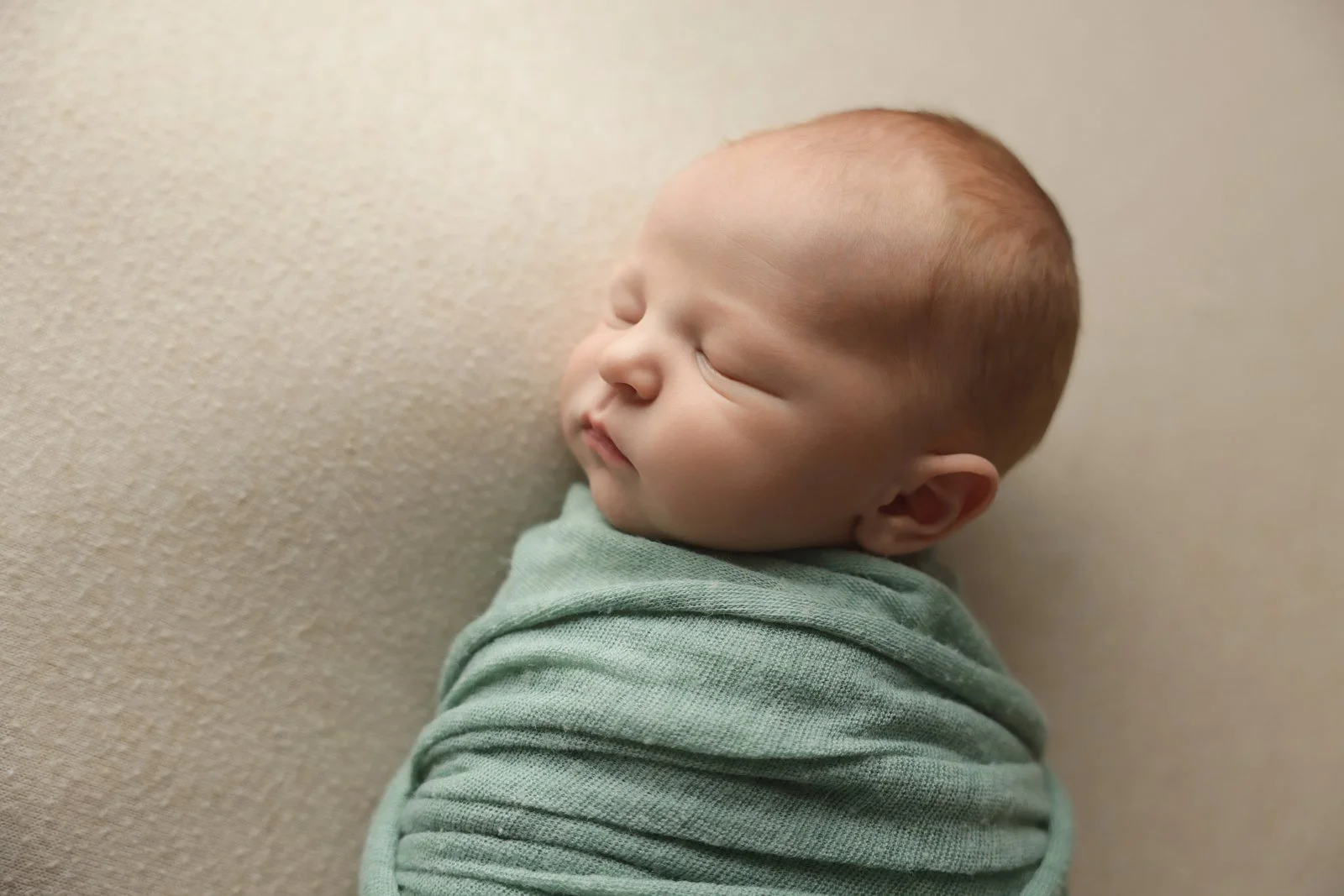 A photo of a newborn baby sleeping with her head turned to the left, wrapped in a sea green blanket on a neutral surface.