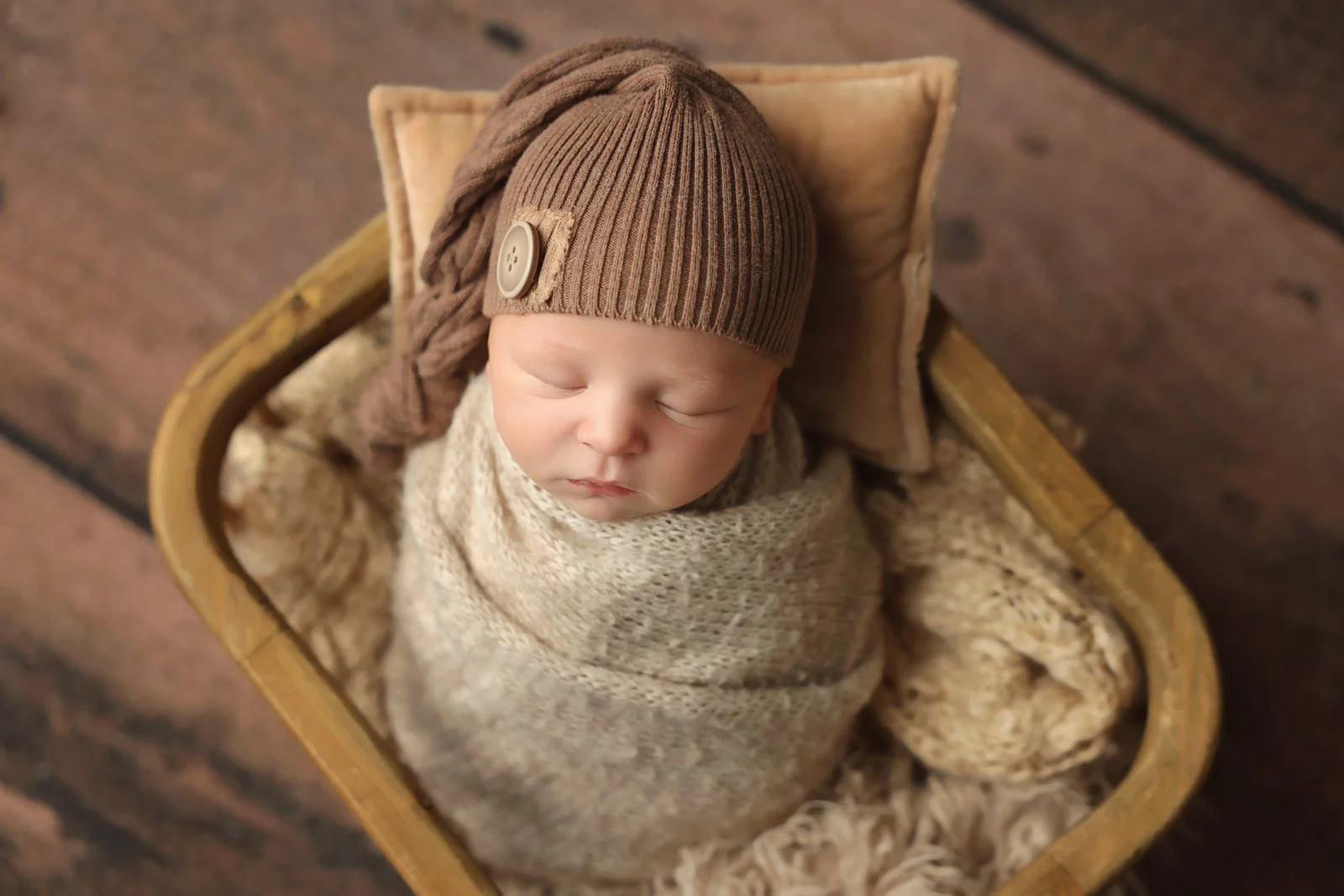 Photo of a newborn baby wrapped in a warm white blanket with a brown hat. She is placed in a wooden box, propped up with a brown pillow, with wood flooring in the background.