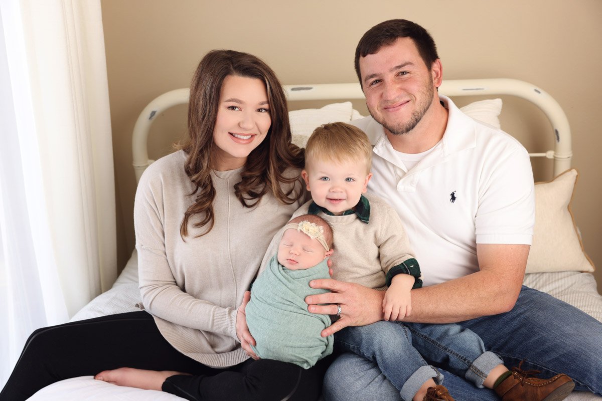 A couple sitting on a metal, twin-sized bed, holding toddler and newborn between them. They are in neutral clothing, and the baby is wrapped in a sea-green blanket. In the background is a light brown wall and a white curtain.