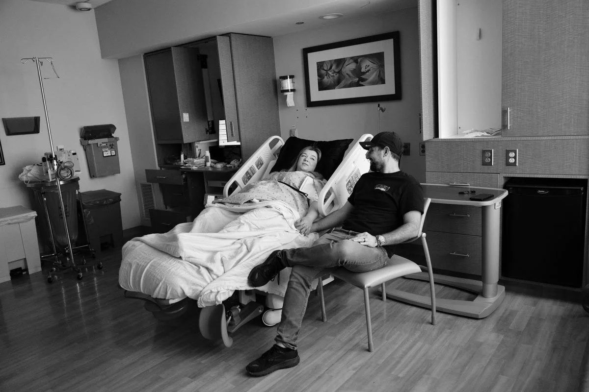 A black and white photo of a woman in a hospital bed holding her husband's hand. Above the bed is a picture frame, and on either side of the bed are cabinets and a variety of hospital equipment.
