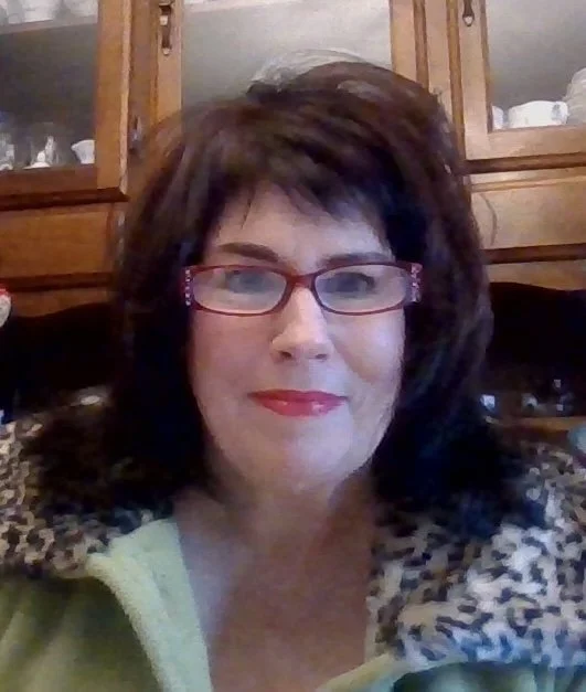 A woman with short dark hair, wearing glasses, red lipstick, and a leopard print jacket, smiling at the camera indoors with wooden cabinets in the background.