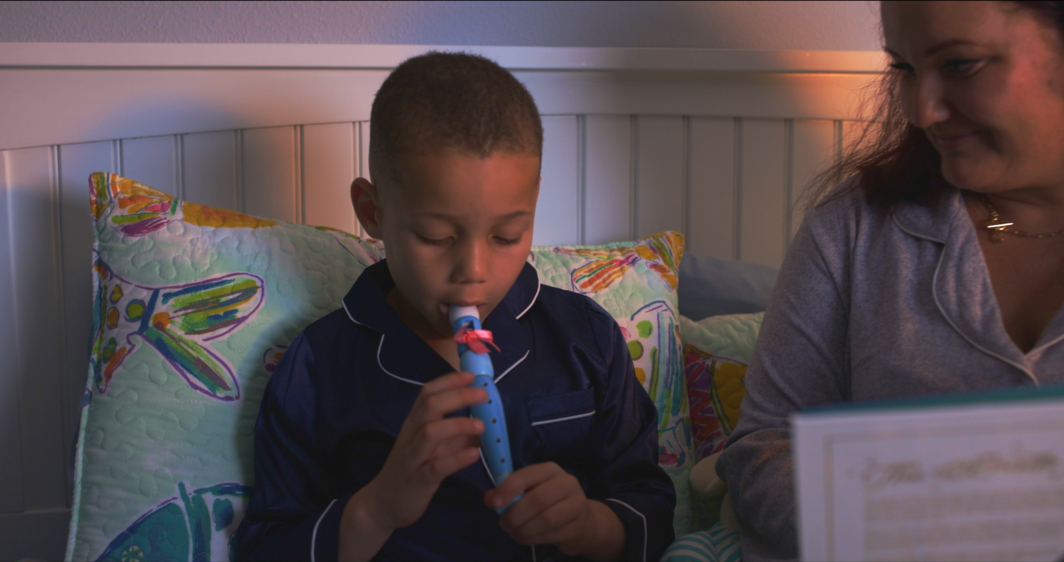A young boy in pajamas sitting in bed playing a the tooth fairy whistle, with his mom beside him looking on.