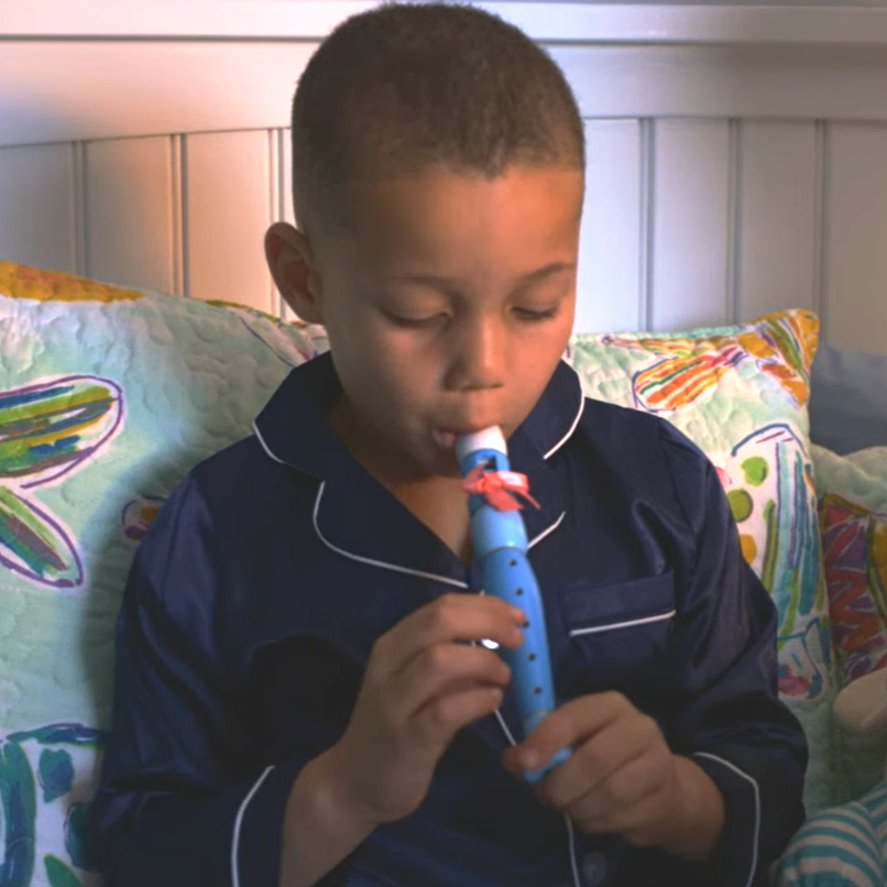 A young boy sitting on a bed with colorful pillows, playing the tooth fairy Bristle Whistle, dressed in dark pajamas with white piping.