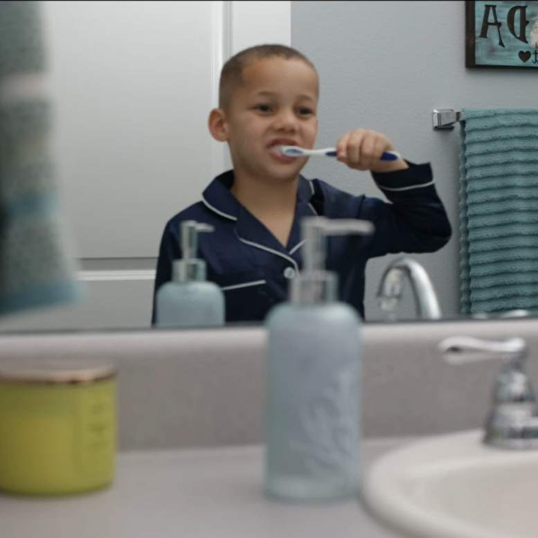 A young boy brushing his teeth while looking at his reflection in the bathroom mirror.
