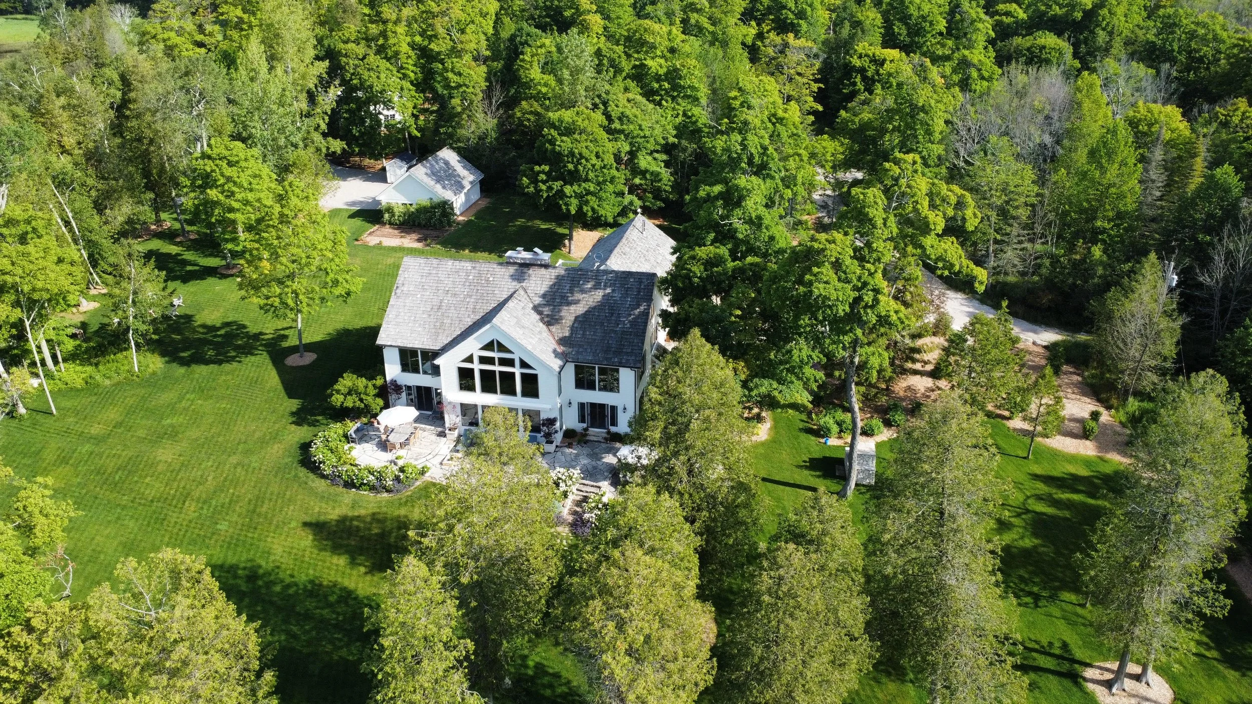 Aerial view of a large house surrounded by lush green trees and a well-maintained lawn, with a driveway and another building nearby.