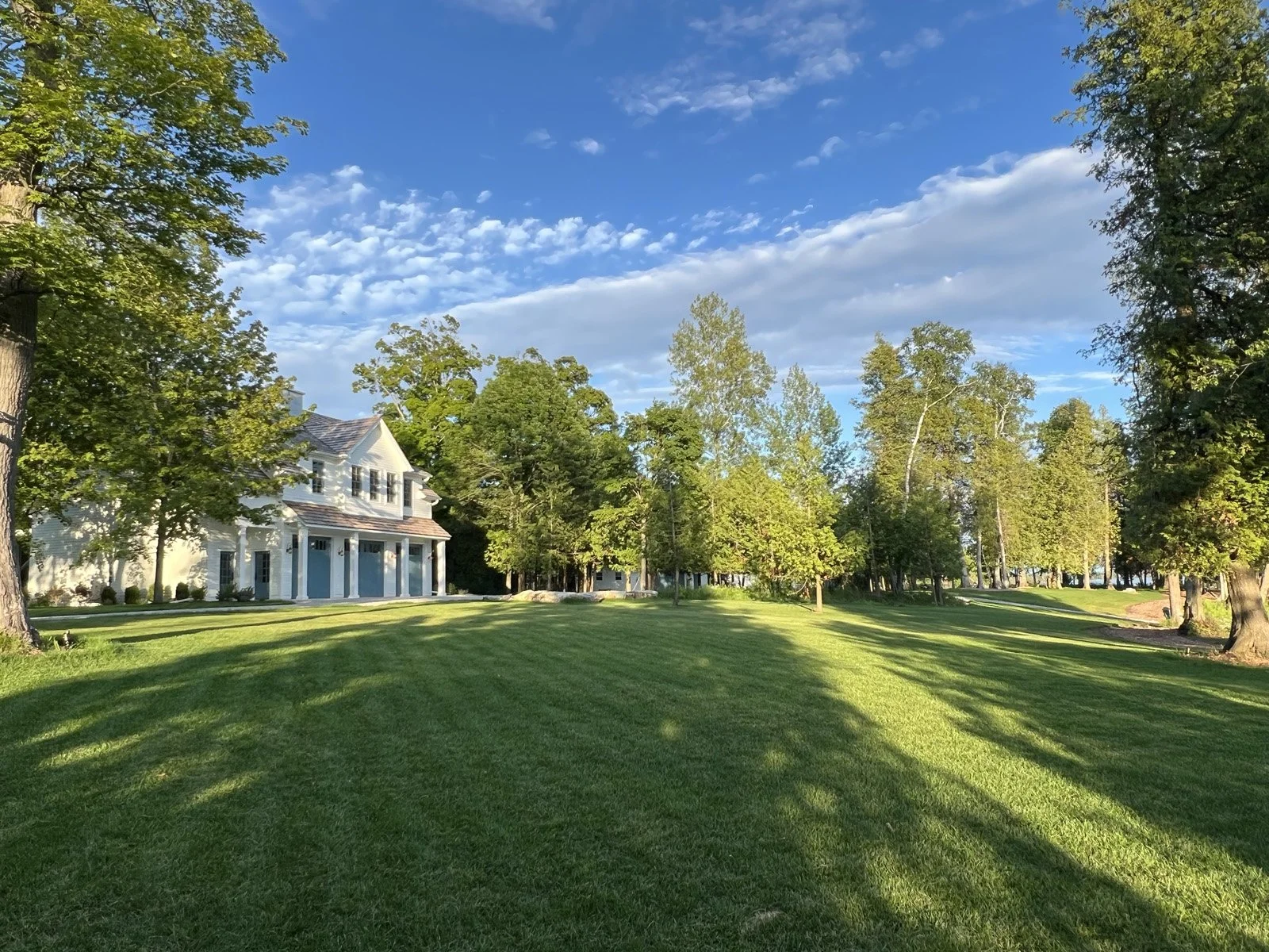 Large white house with a wraparound porch on a sunny day, surrounded by lush green lawn and tall trees.