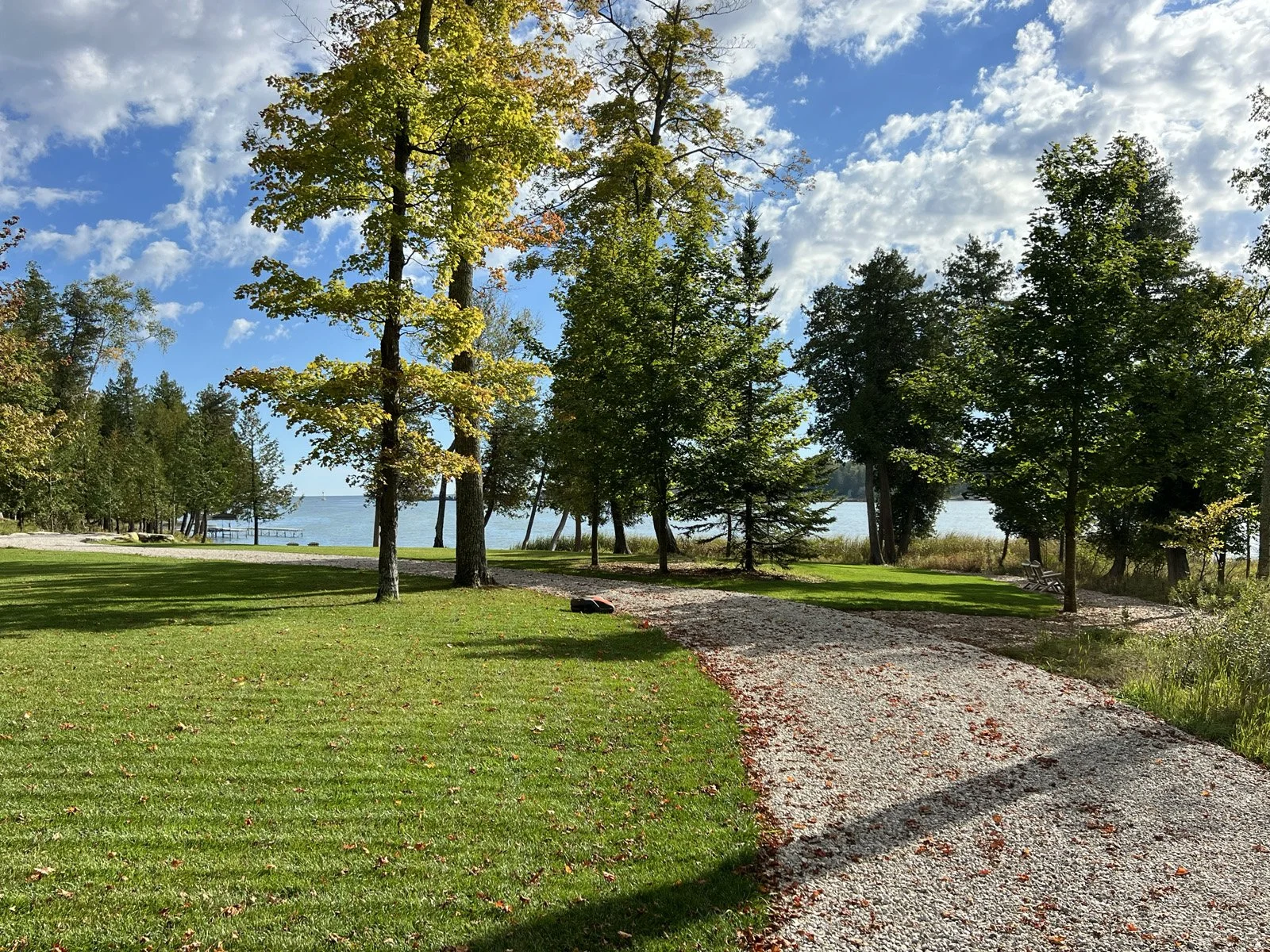 Scenic view of a grassy area with trees, a gravel path, and a glimpse of a lake in the background under a partly cloudy sky.