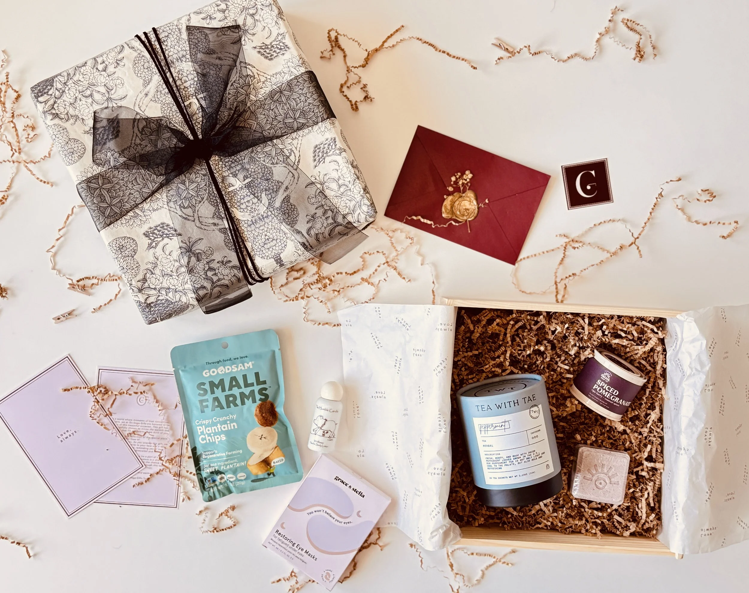 Flat lay of a gift box with a black ribbon, a red envelope with a wax seal, a small jar, a blue tea can, a pink soap, a small bottle, a pack of plantain chips, and two cards, surrounded by decorative shredded paper.
