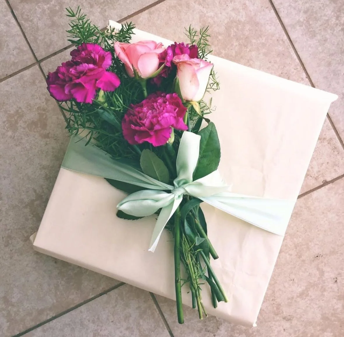 A bouquet of pink and purple flowers with green leaves tied with a white ribbon, placed on a wrapped white gift box on a tiled floor.