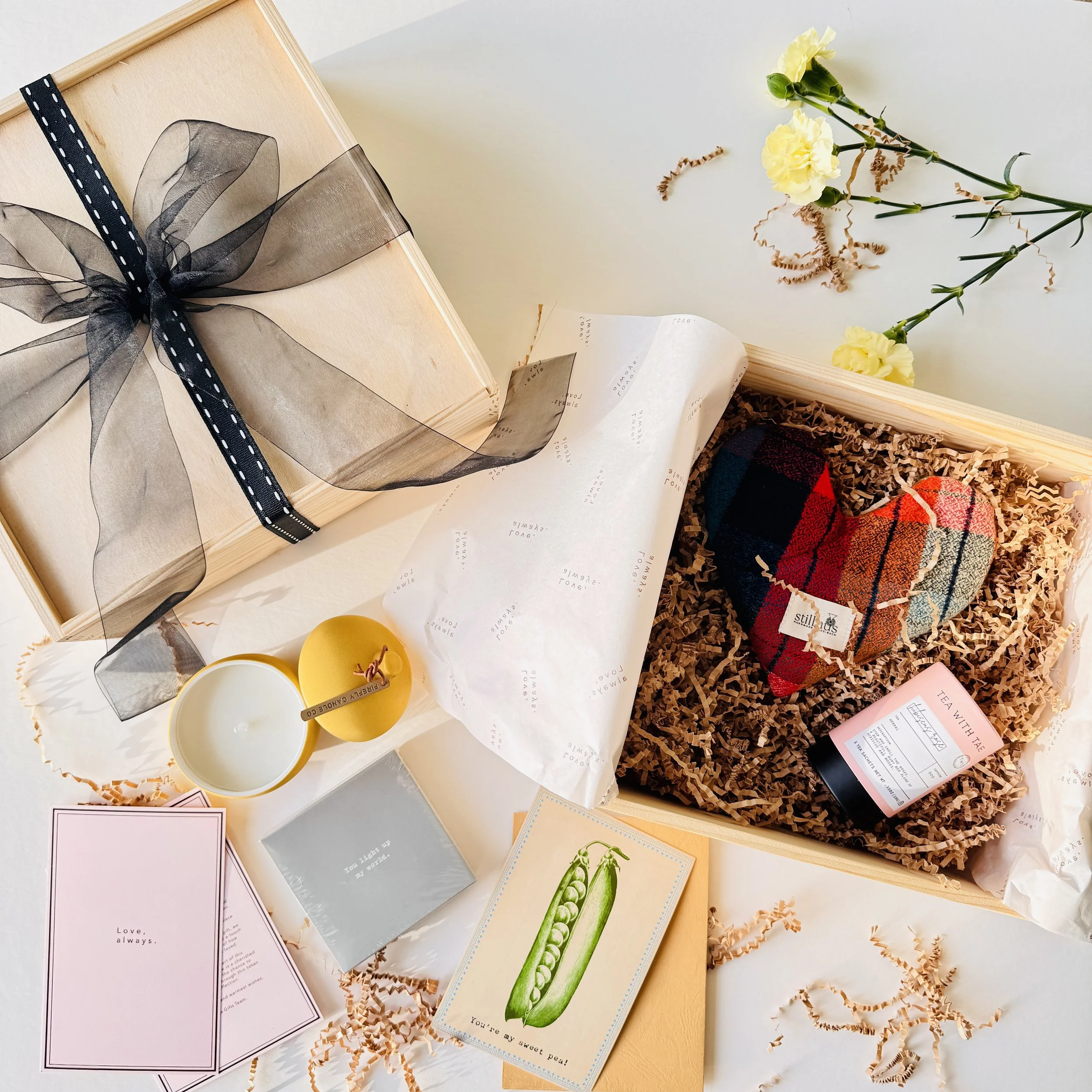 A gift box with a black ribbon and bow, containing a tartan plush heart and a tube of tea, surrounded by a open pink card, a yellow candle, a small envelope, a postcard with a pea pod illustration, and yellow flowers, all on a white surface with crinkled tissue paper.