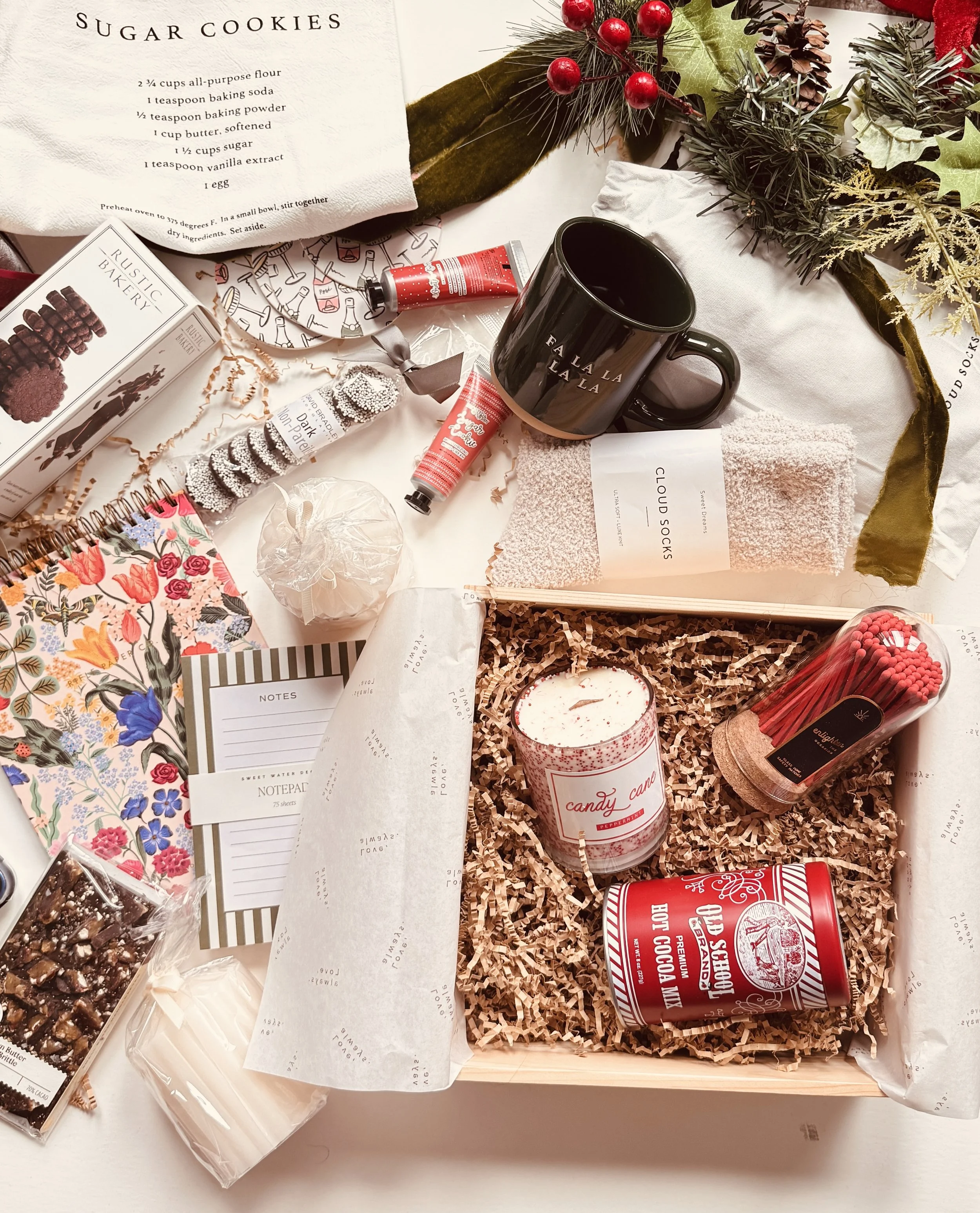 Holiday gift basket with candles, snacks, holiday-themed items, a mug, socks, a notebook, and festive decorations on a white table.