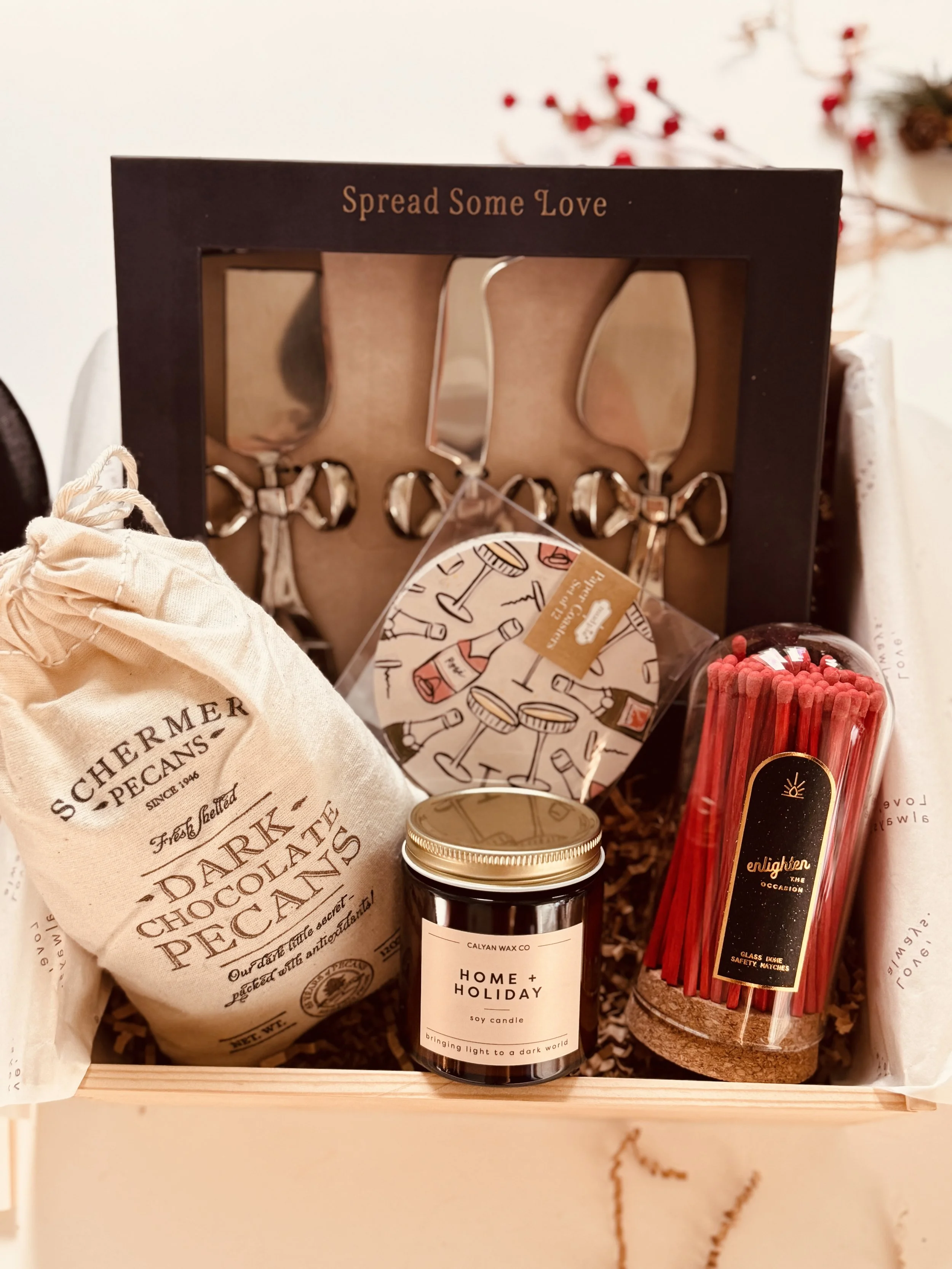 A gift box containing a bag of Schermer pecans, a soy candle labeled 'Home + Holiday,' a set of three small spreading knives, a container of red matches, and a small box with illustrated wine and drink glasses, with a frame at the back that says 'Spr