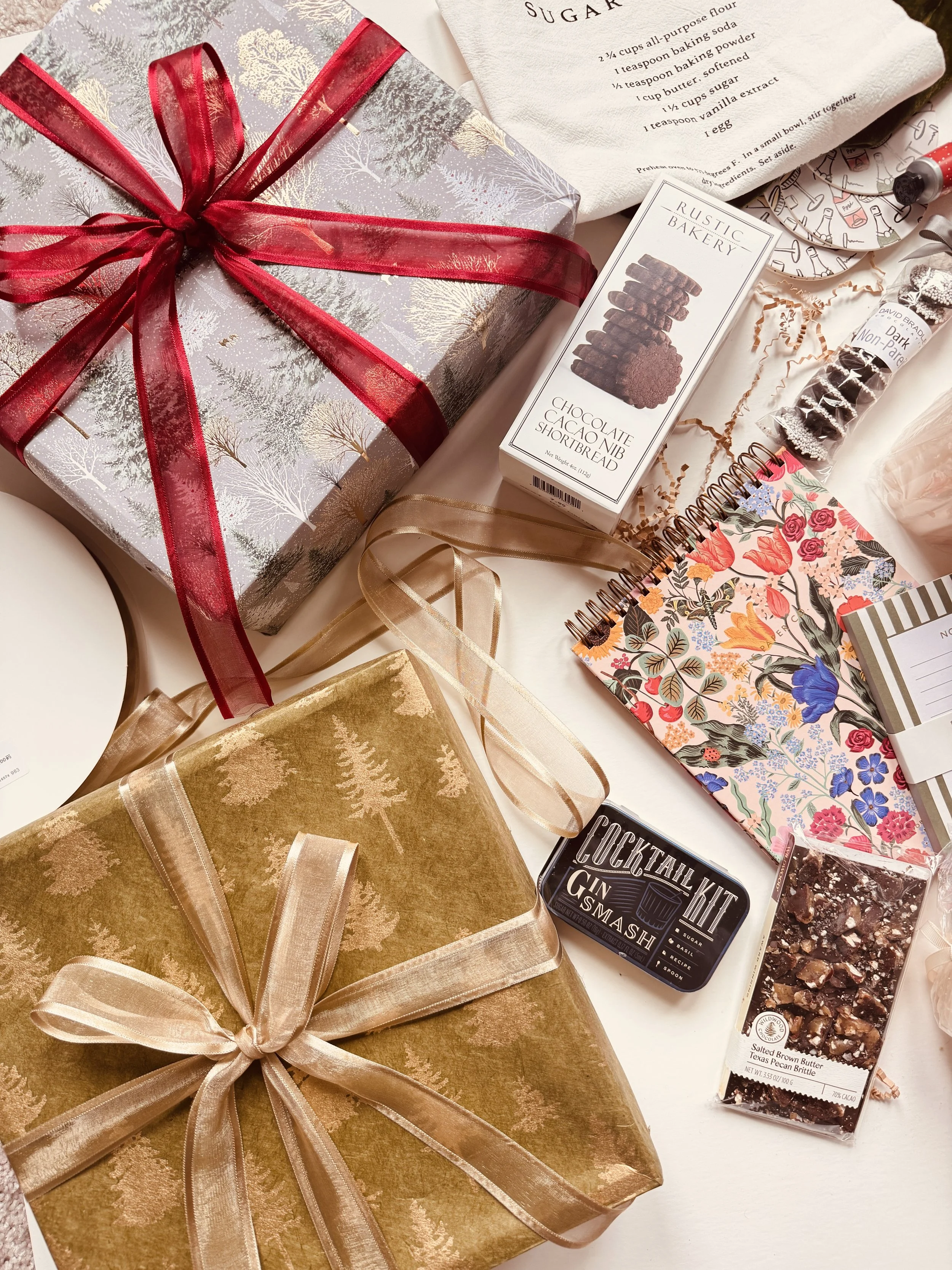 Arranged holiday gifts and treats, including wrapped presents with ribbons, a box of chocolate cookies, baking supplies, a spiral notebook with a floral cover, and a tin labeled 'Cocktail Kit Gin Smashes.'