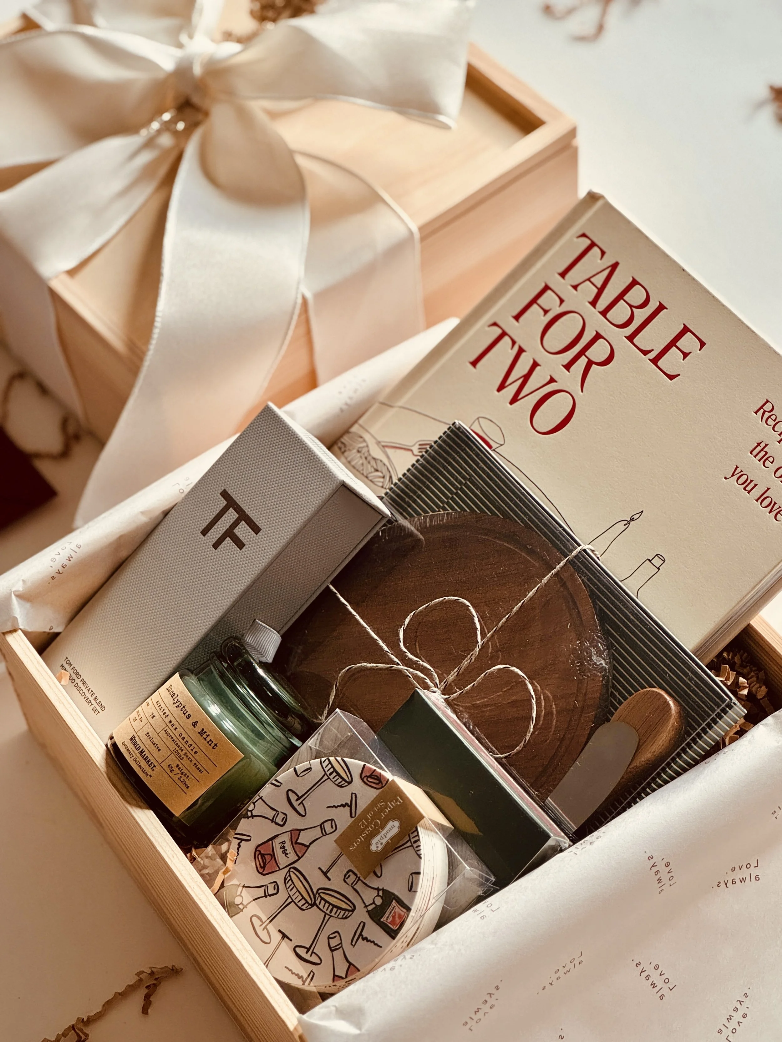 A gift box with a ceramic mug, a book titled 'Table for Two,' a candle, a small bottle, and a round coaster with cocktail illustrations.