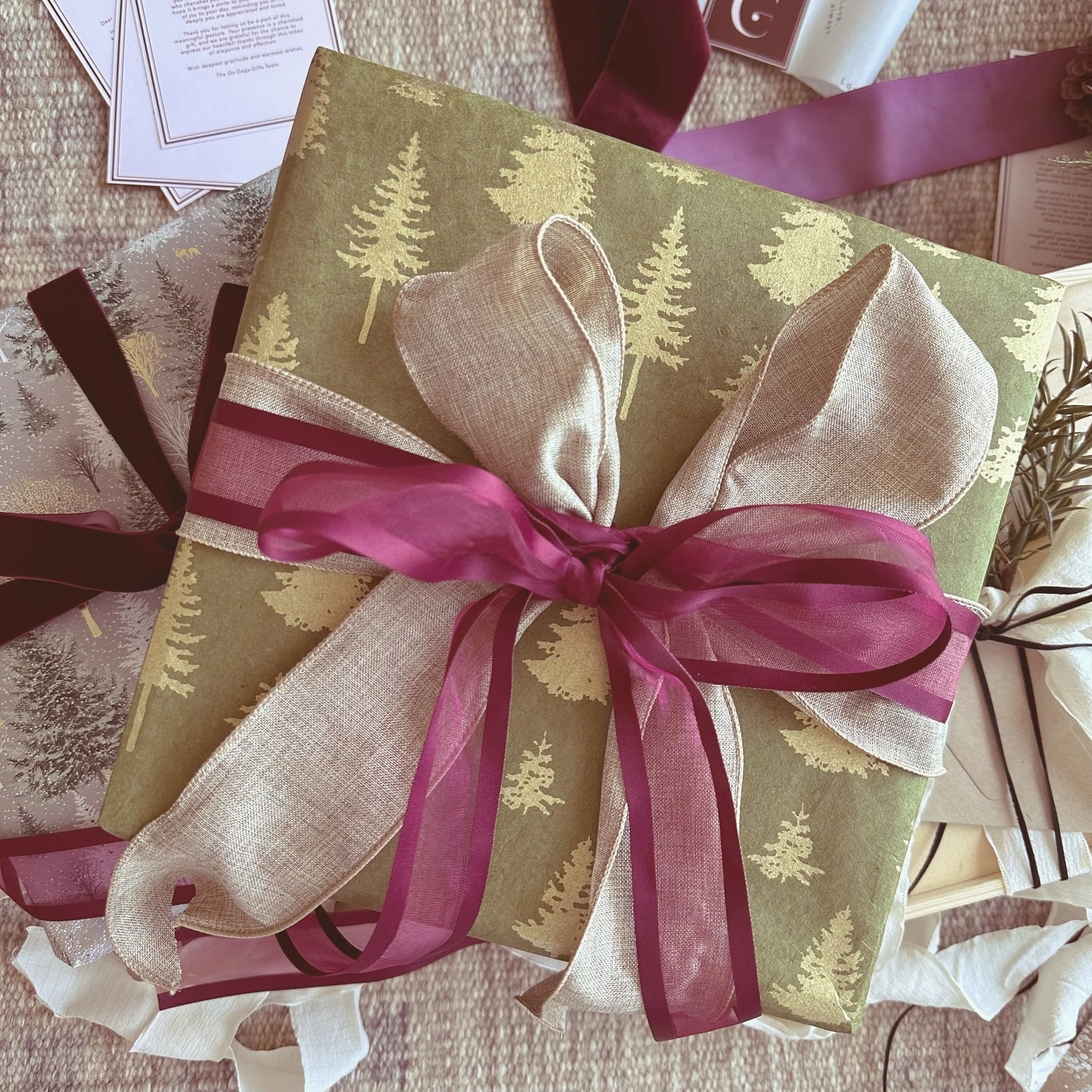 A wrapped Christmas gift with green paper featuring gold Christmas trees, beige ribbon, and a maroon sheer ribbon bow. Surrounding the gift are other wrapped presents, papers, and ribbons on a beige carpet.