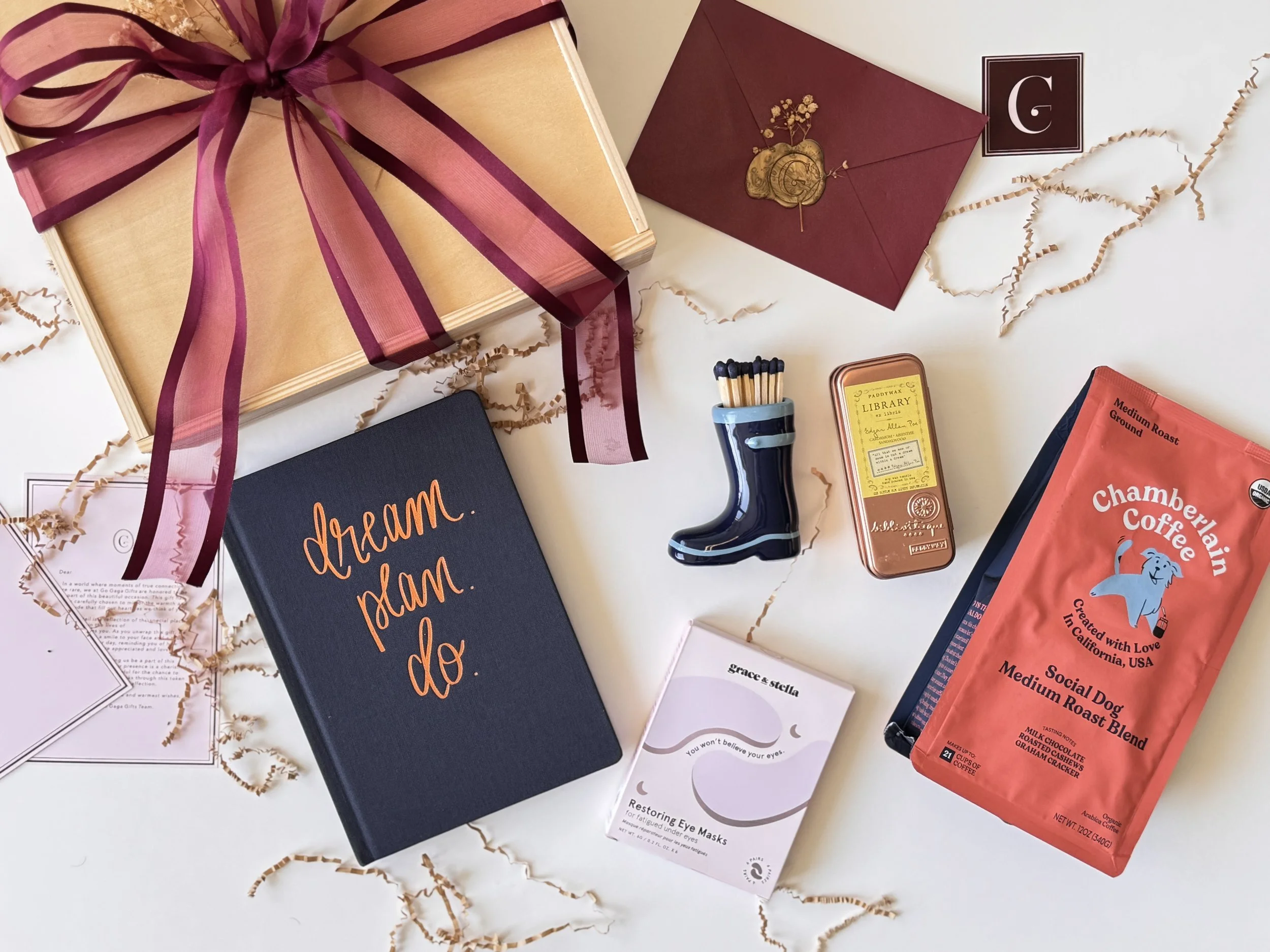 Flat lay of assorted gift items, including a wrapped box with a pink ribbon, a red envelope, a sock-shaped container of matches, a tin of library mints, a bag of coffee, a small eye mask box, a navy notebook with orange writing, and some shredded pap