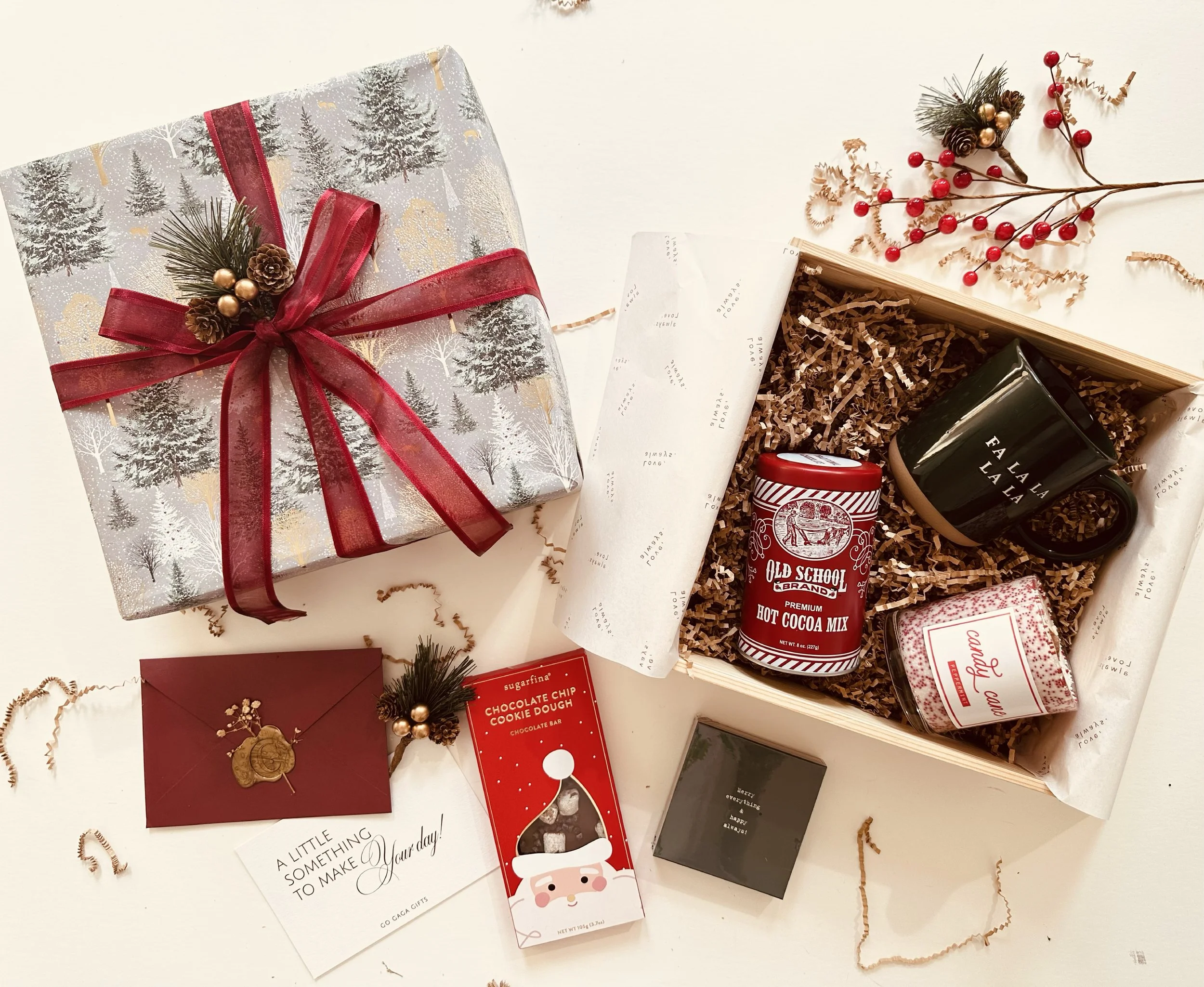 Christmas gift box wrapped in festive paper with a red ribbon and holiday decorations, an open gift box containing holiday-themed mugs, a hot cocoa mix, a candle, and holiday cards, surrounded by decorative sprigs and paper shreddings.