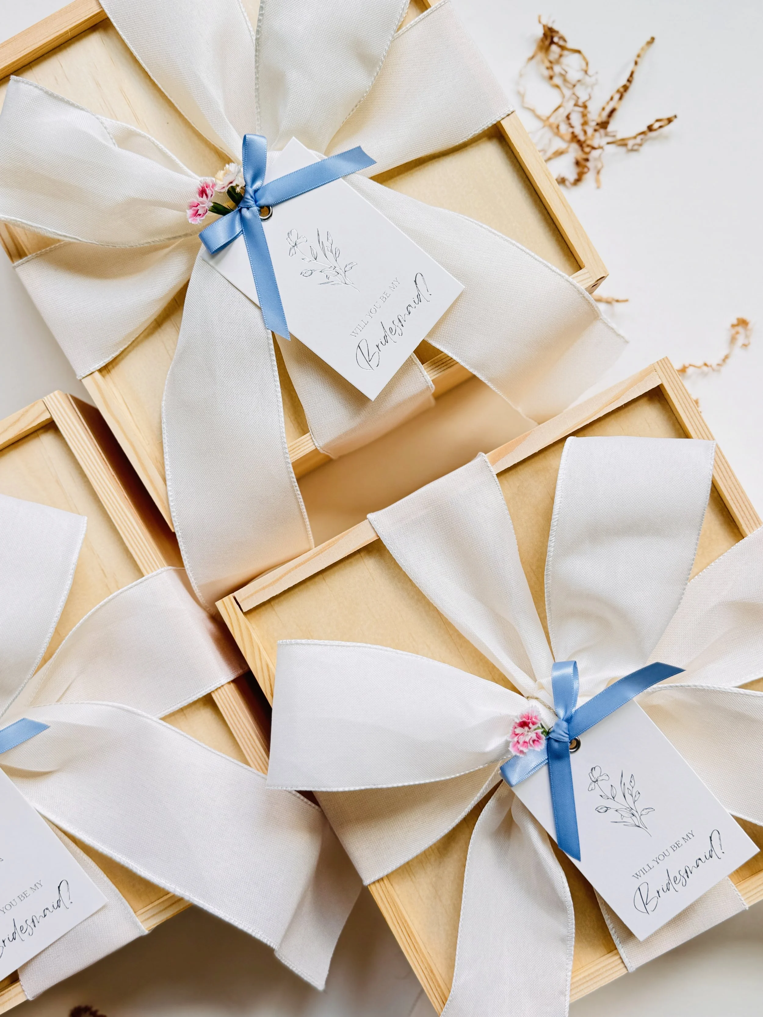 Luxury bridesmaid proposal gift boxes in pinewood packaging, tied with white silk ribbons, blue bows, and personalized floral tags.