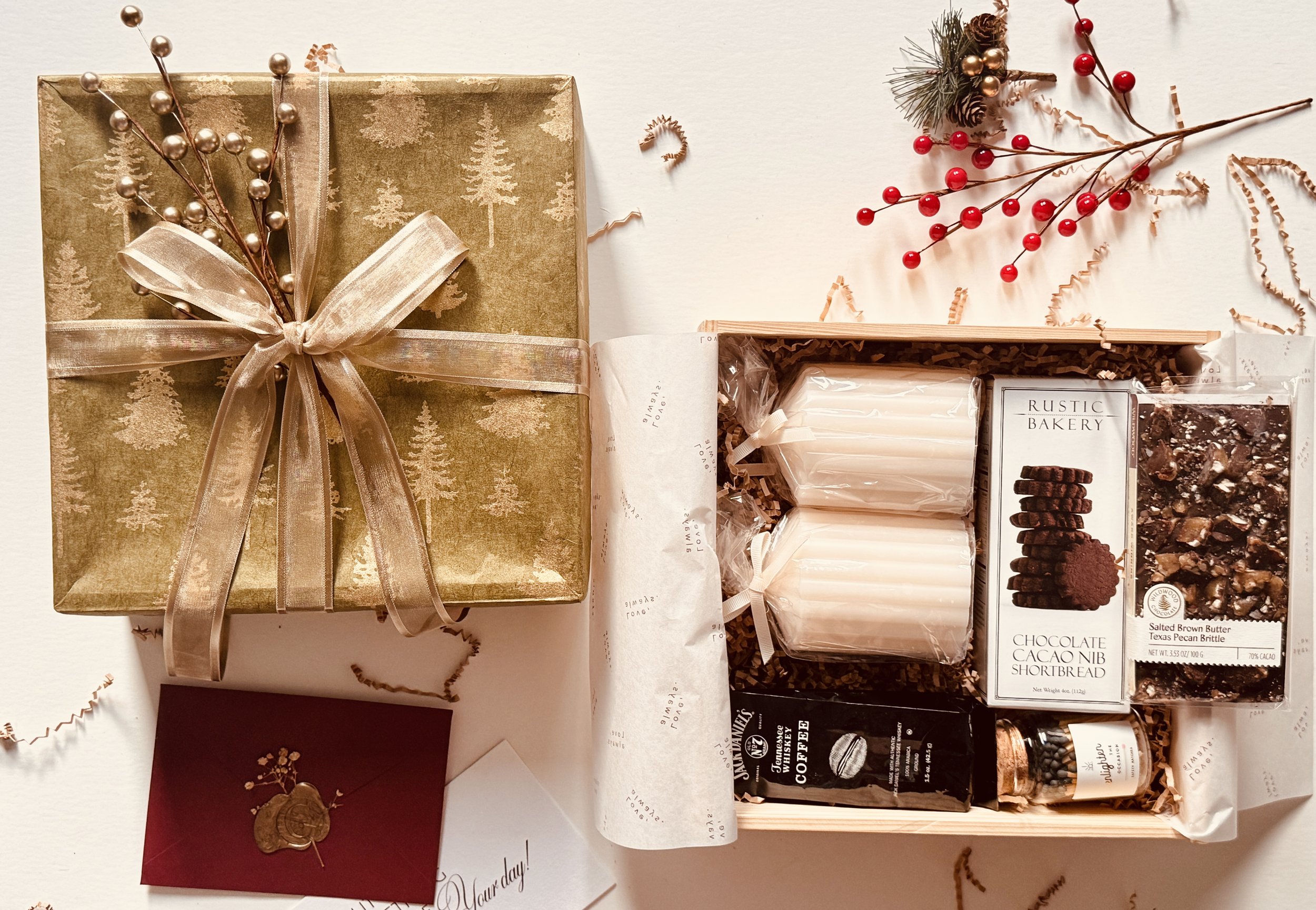 A Christmas gift box with gold wrapping paper decorated with pine tree patterns and a champagne-colored ribbon bow, surrounded by holiday decorations, and an open gift box containing chocolates, marshmallows, coffee, and other treats.