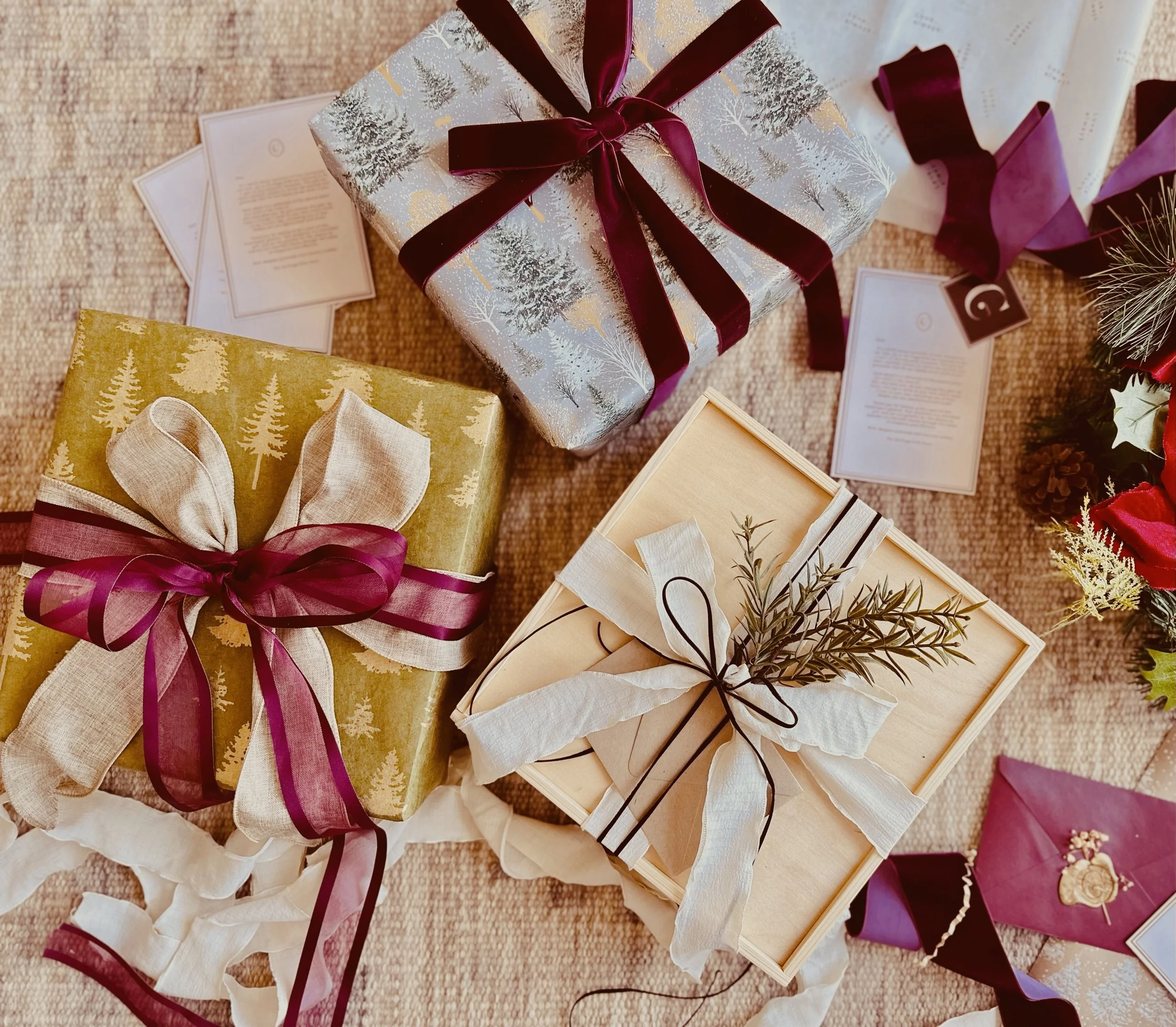 Flatlay of holiday gift boxes wrapped in gold, silver, and wooden packaging with elegant burgundy and cream ribbons, dried greenery, festive envelopes, and holiday cards styled on a neutral textured background.