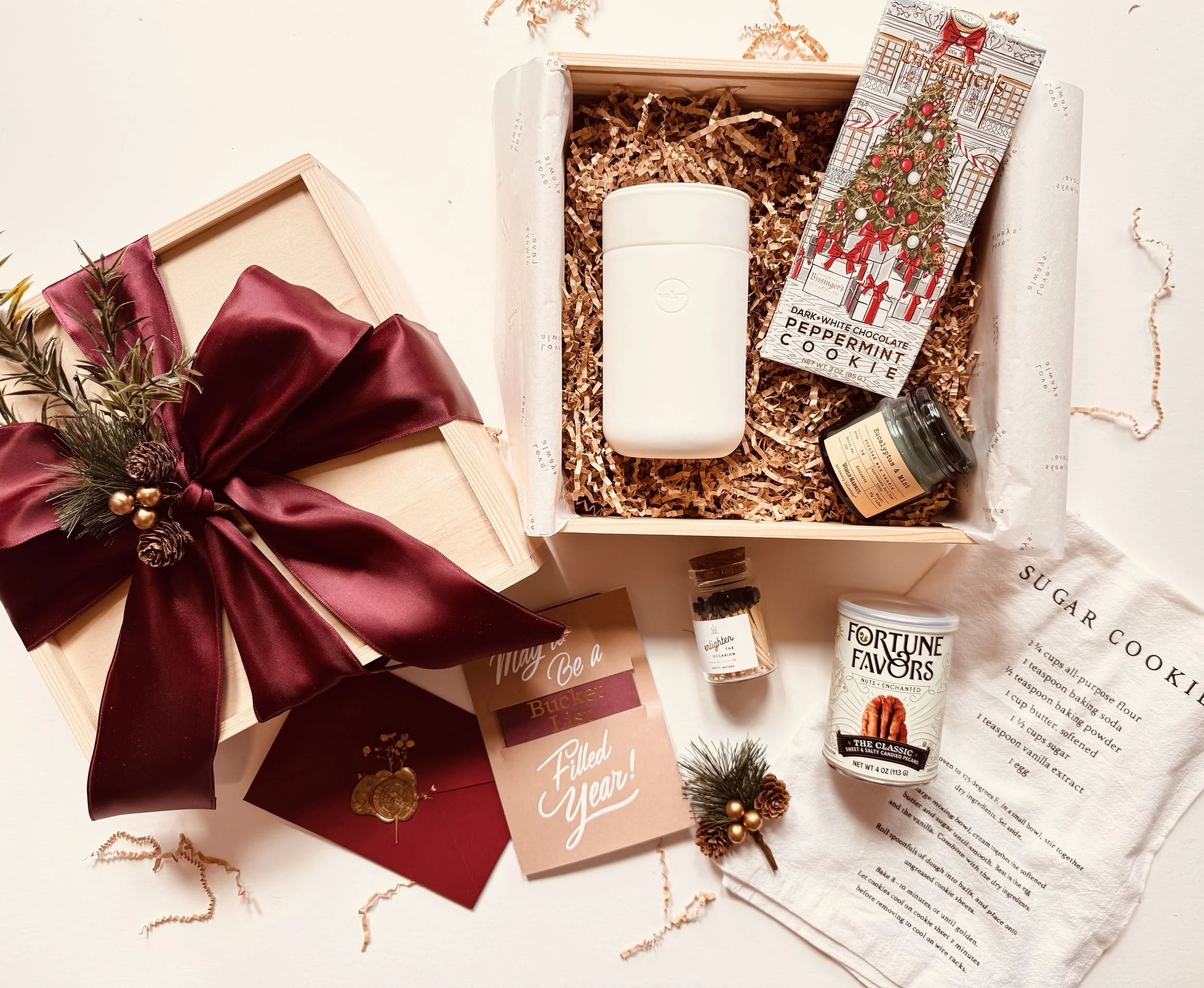 A holiday gift box with a burgundy satin ribbon, Christmas decorations, and gourmet food items, including a box of dark white chocolate peppermint cookies, a jar of chocolate spread, a tin of fortune favors nuts, and a spice jar. There is also a holi