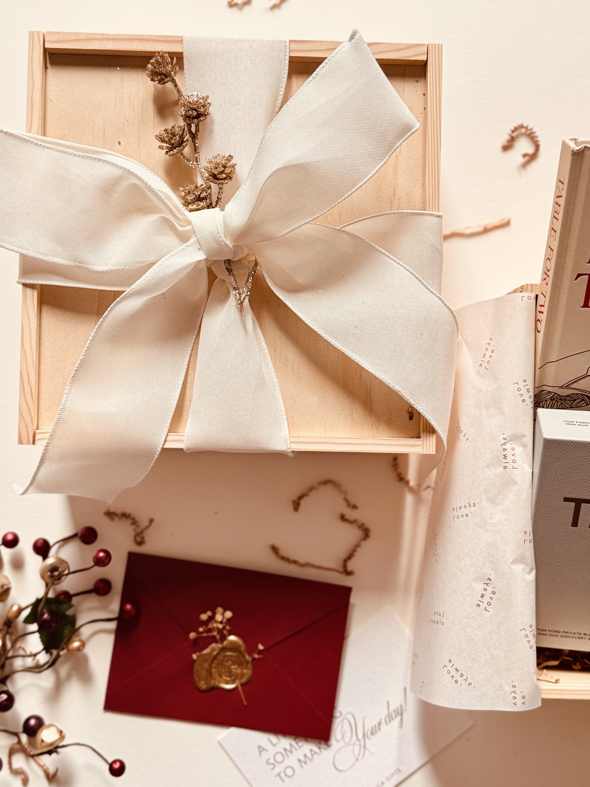 Open gift box with a large cream-colored satin bow, decorative sprig, and jewelry inside. Gift paper and other items surrounding the box.
