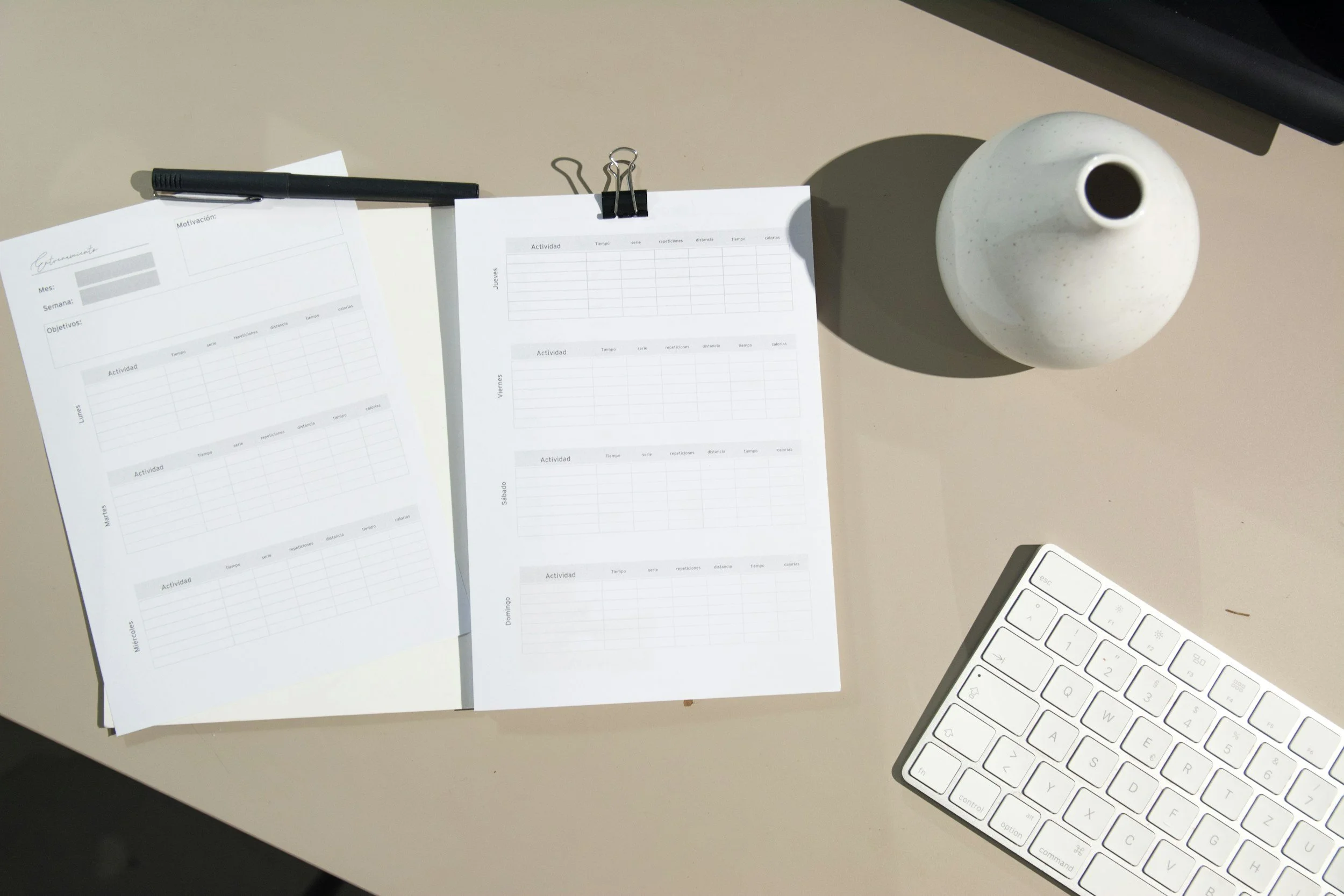 A beige office desk with two sheets of paper containing tables, a black pen, and a white keyboard.