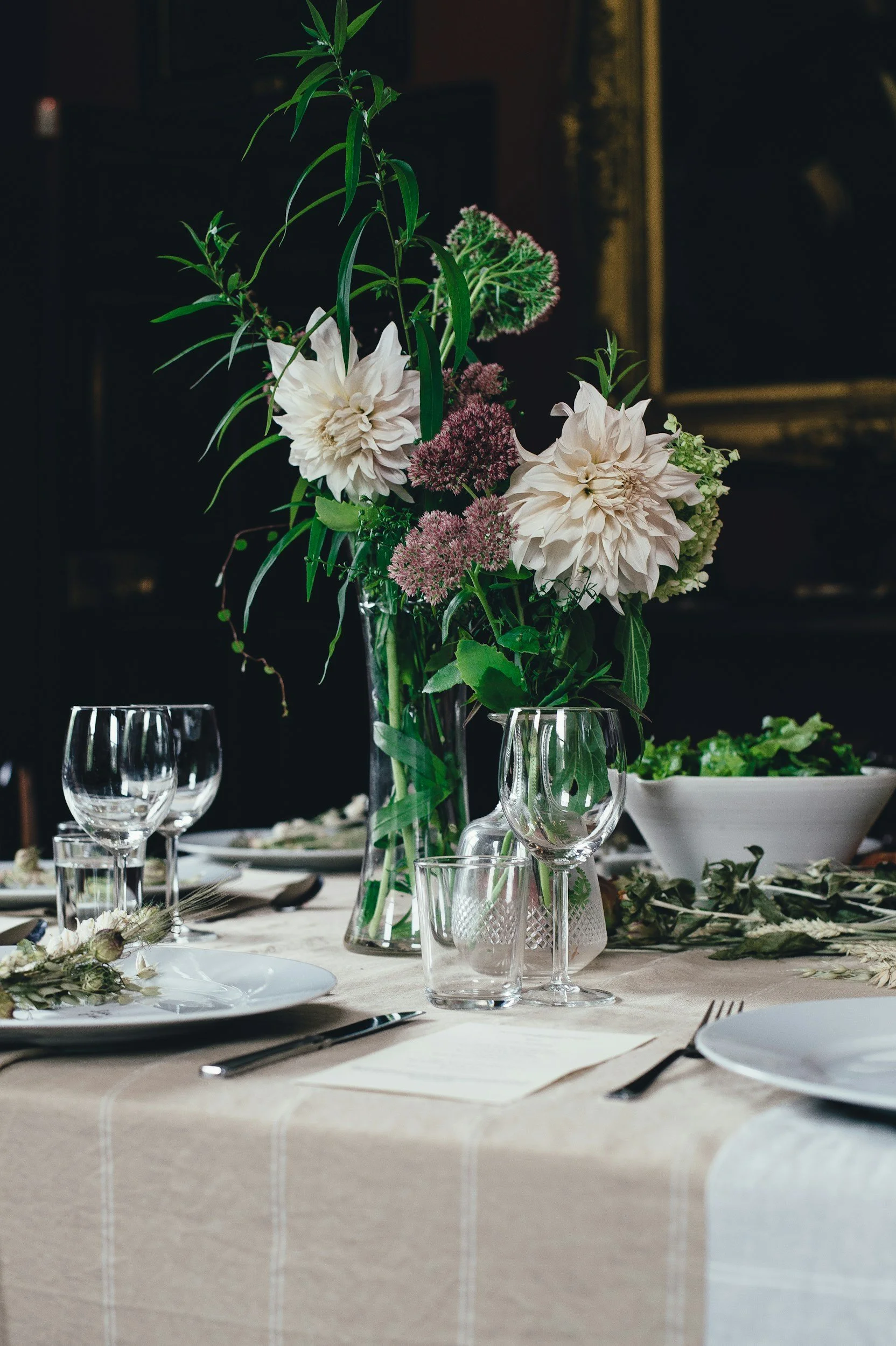 A table set for a formal dinner with a tall glass vase filled with a bouquet of white dahlias, purple alliums, and greenery. The table has white plates, clear glasses, silverware, and a beige tablecloth with a subtle checkered pattern. In the background, there are wine glasses and a salad bowl, with a dark, out-of-focus setting.