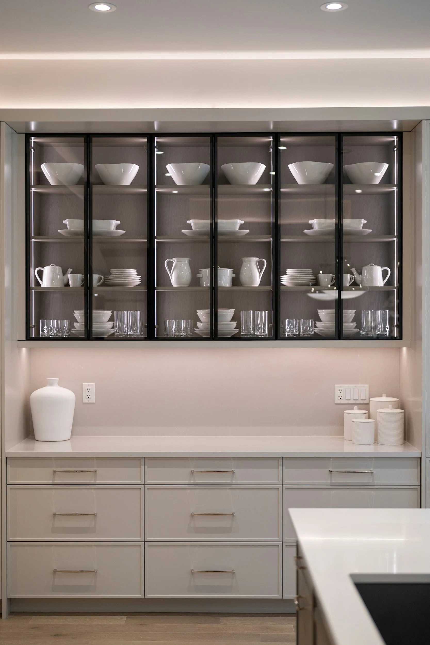 A modern kitchen cabinet filled with white dishware including bowls, plates, pitchers, and cups, with a large white vase on the countertop and a kitchen counter in the foreground.