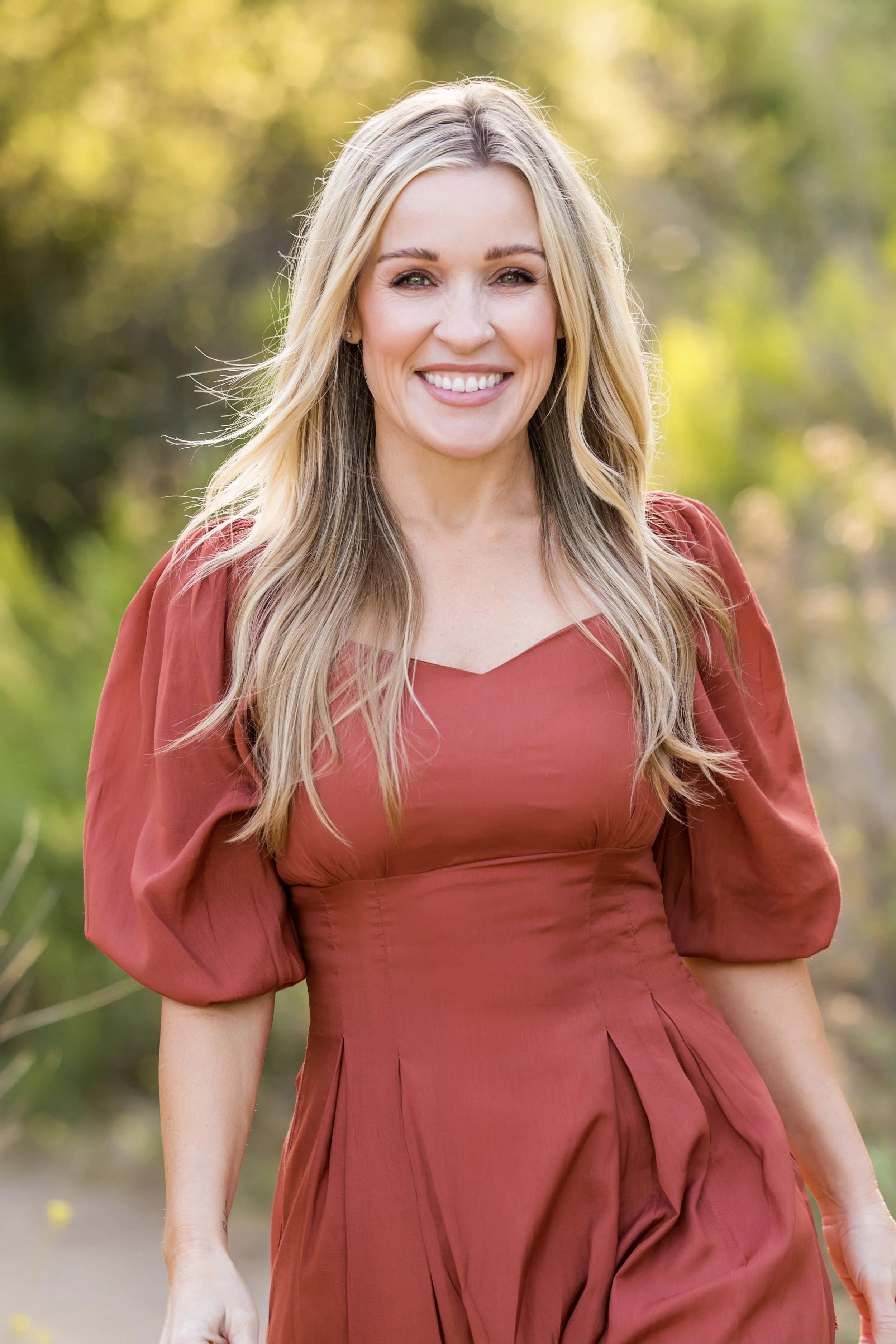 A woman with long blonde hair wearing a red dress smiling outdoors with a blurred natural background.