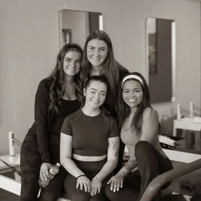 Four women smiling together in a room with mirrors and a television mounted on the wall.